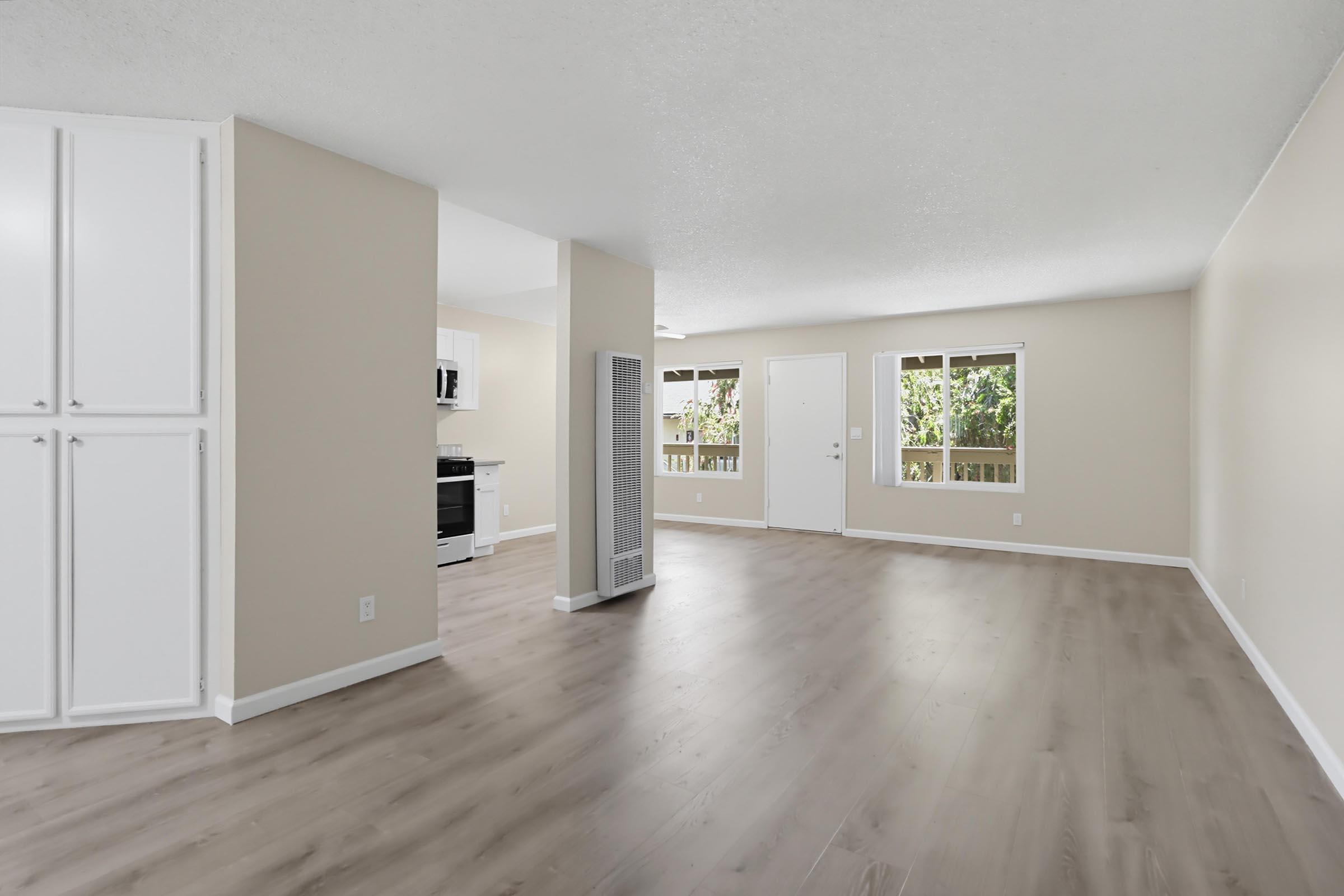 A spacious, empty living area with light-colored walls and laminate flooring. It features two windows allowing natural light, a door leading outside, and a small kitchen area in the background with a stove and microwave.