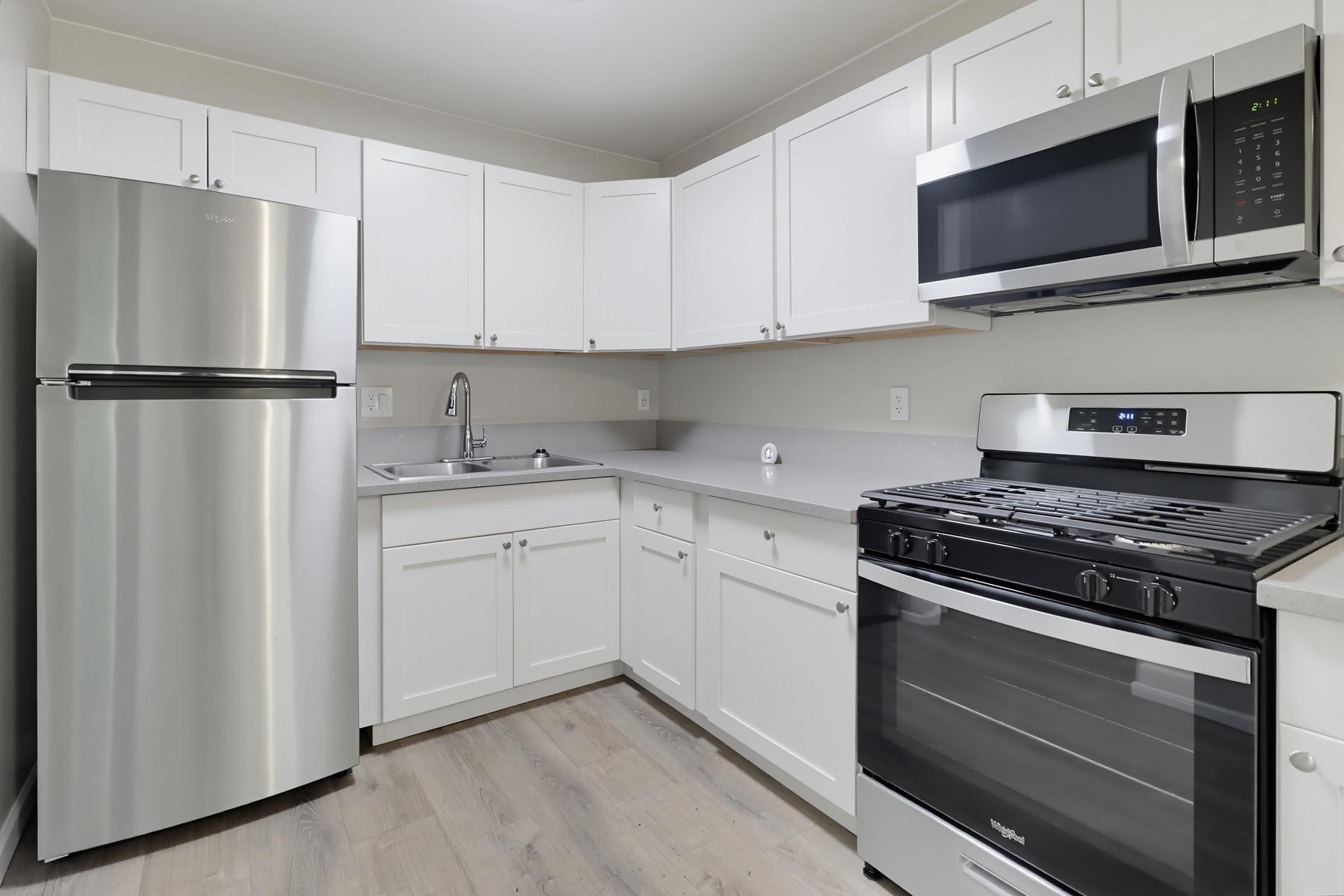 A modern kitchen featuring white cabinetry, a stainless steel refrigerator, a stainless steel gas range, and a built-in microwave. The countertop is light gray, and there is a sink with a faucet. The flooring is a light wood laminate, creating a bright and clean atmosphere.
