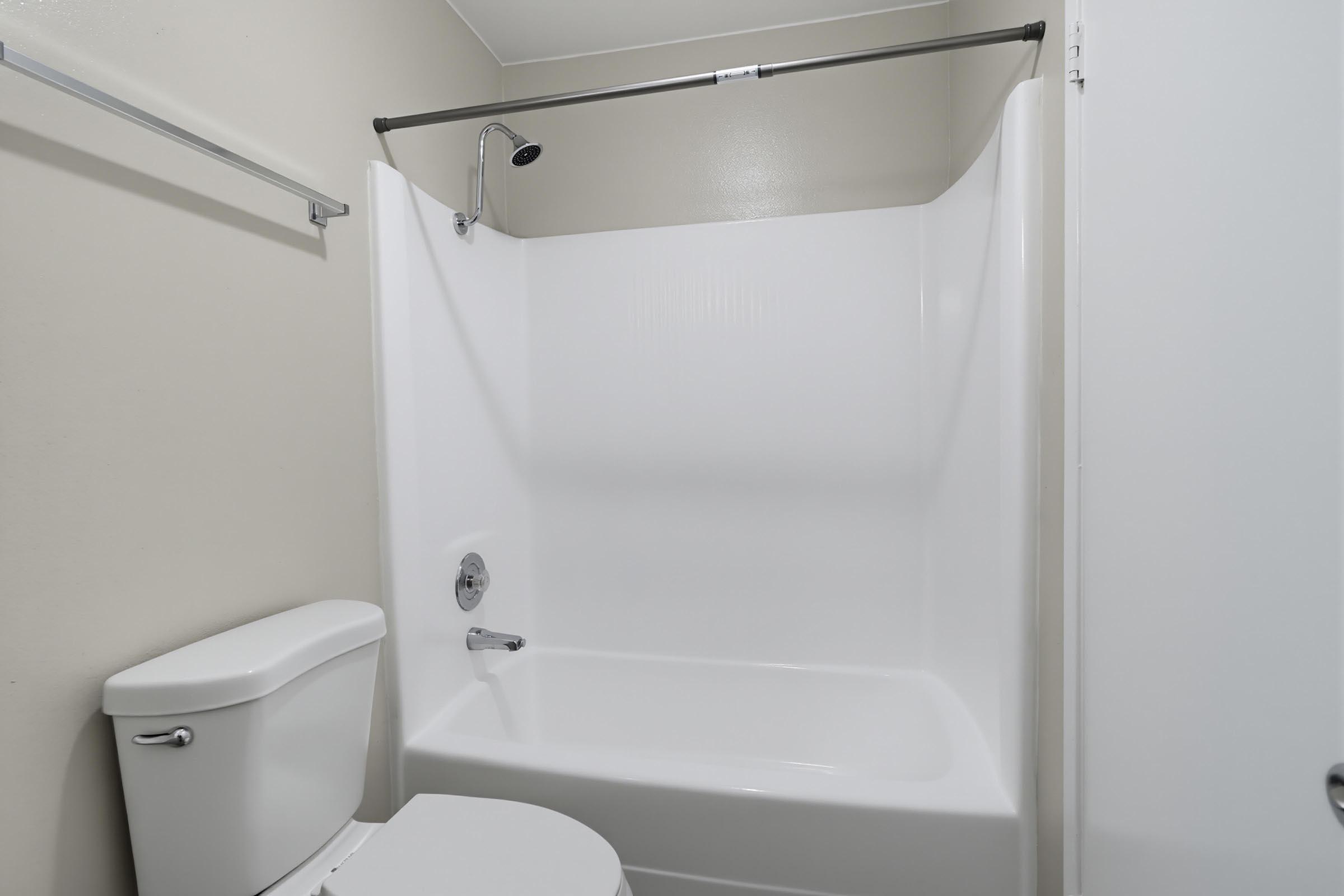 A clean and modern bathroom featuring a white bathtub and shower, a toilet, and light-colored walls. The shower has a simple chrome showerhead and a shower curtain rod. The overall design is minimalistic and well-maintained.