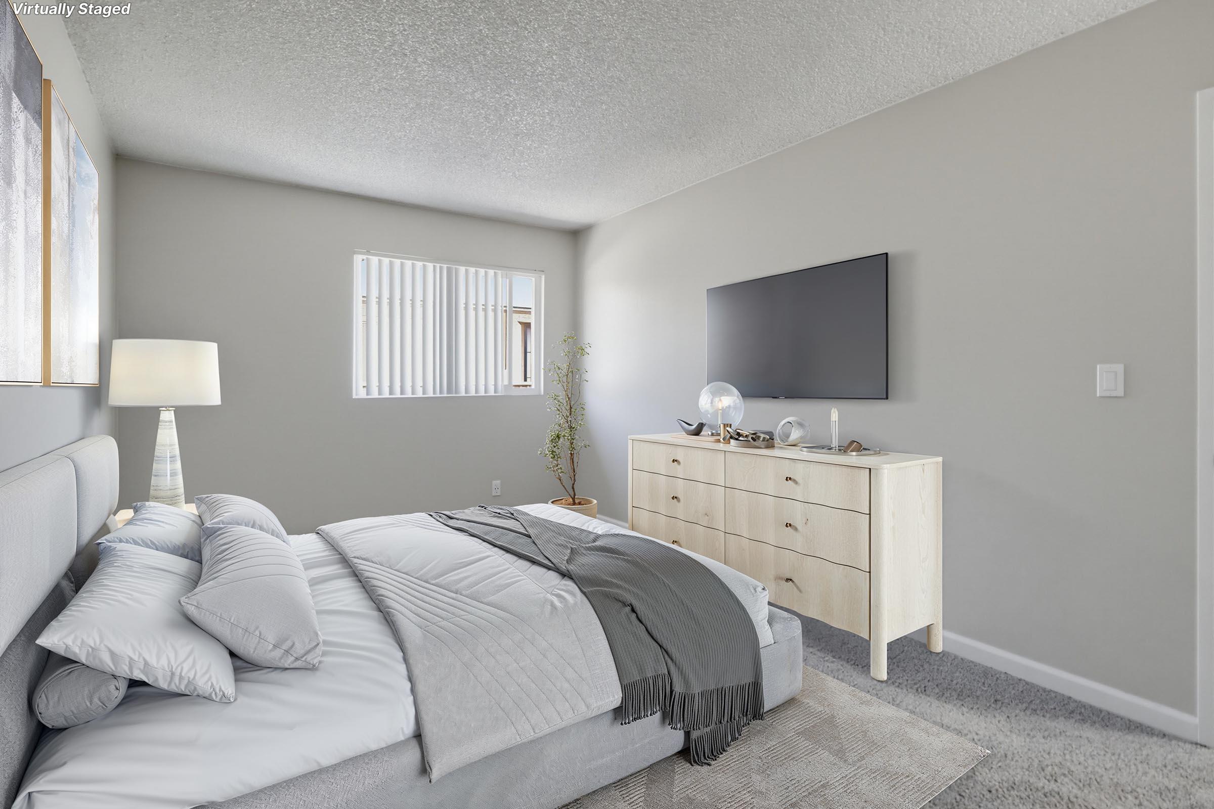 A modern bedroom featuring a light gray bed with soft bedding and decorative pillows. There is a nightstand with a lamp, a light-colored dresser with decorative items, and a wall-mounted flat-screen TV. The room has neutral-colored walls and a window with blinds, creating a bright and airy atmosphere.
