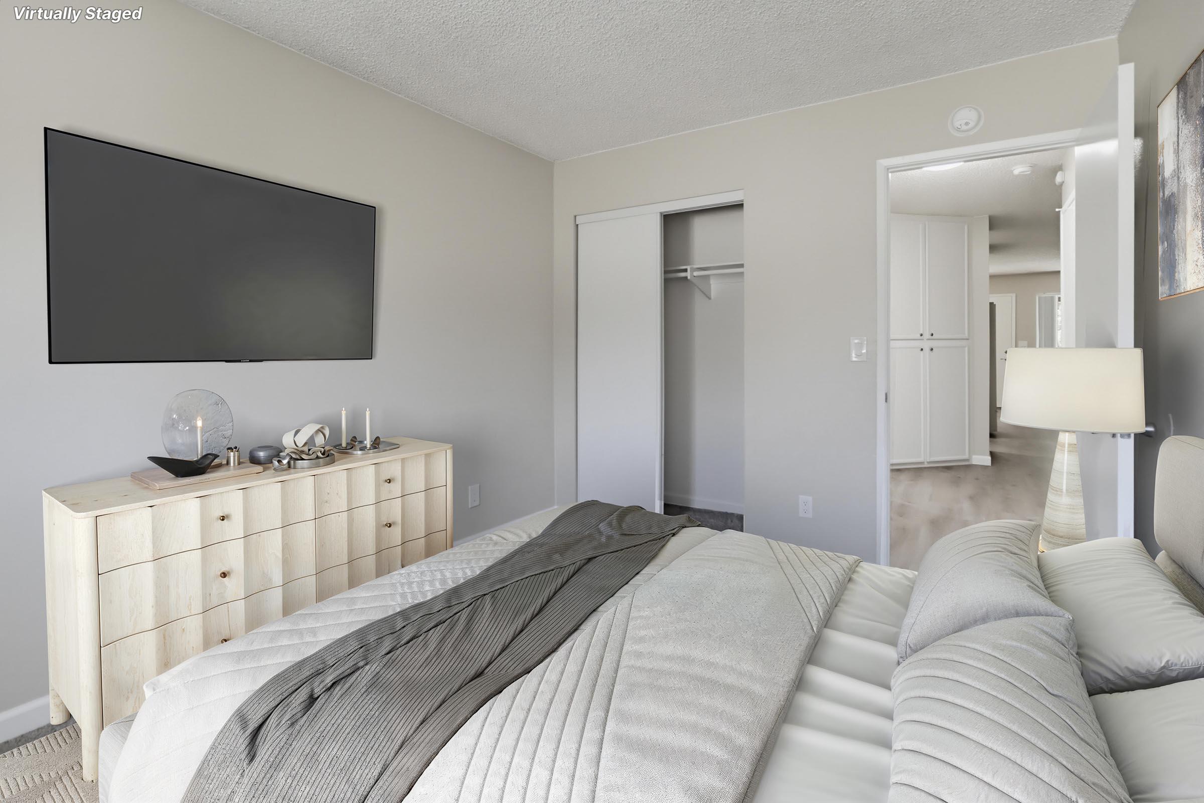 A modern bedroom featuring a neatly made bed with gray bedding and a matching throw, a stylish dresser with decorative items, and a wall-mounted television. A closet door is visible, while a doorway leads to another room. The walls are painted in neutral tones, creating a bright and inviting atmosphere.