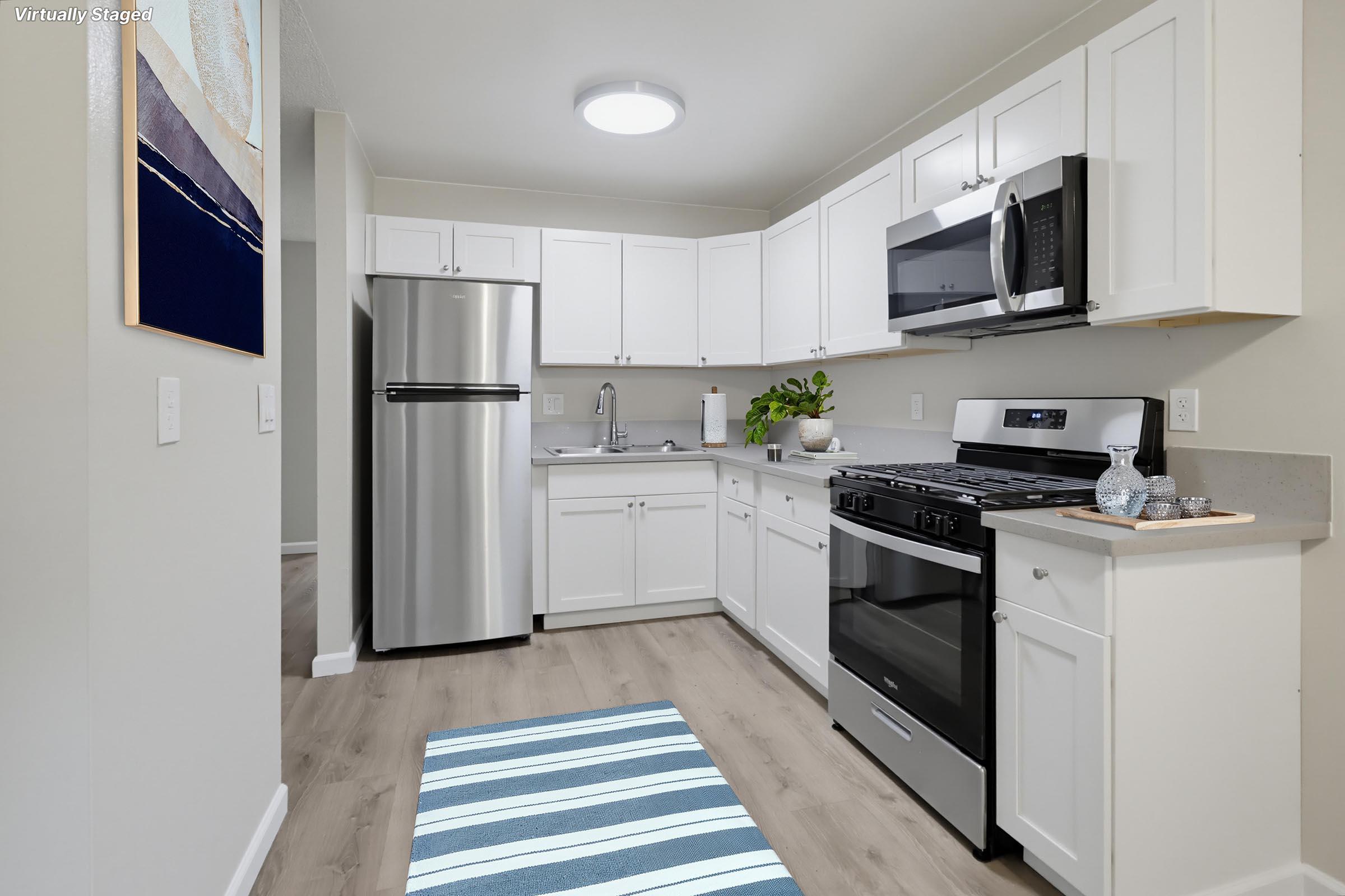 A modern kitchen featuring white cabinetry, stainless steel appliances including a refrigerator and microwave, and a gas stove. The countertops are light-colored with a gray backsplash. A blue and white striped rug is on the floor, with a small plant and kitchen items on the countertop, illuminated by overhead lighting.