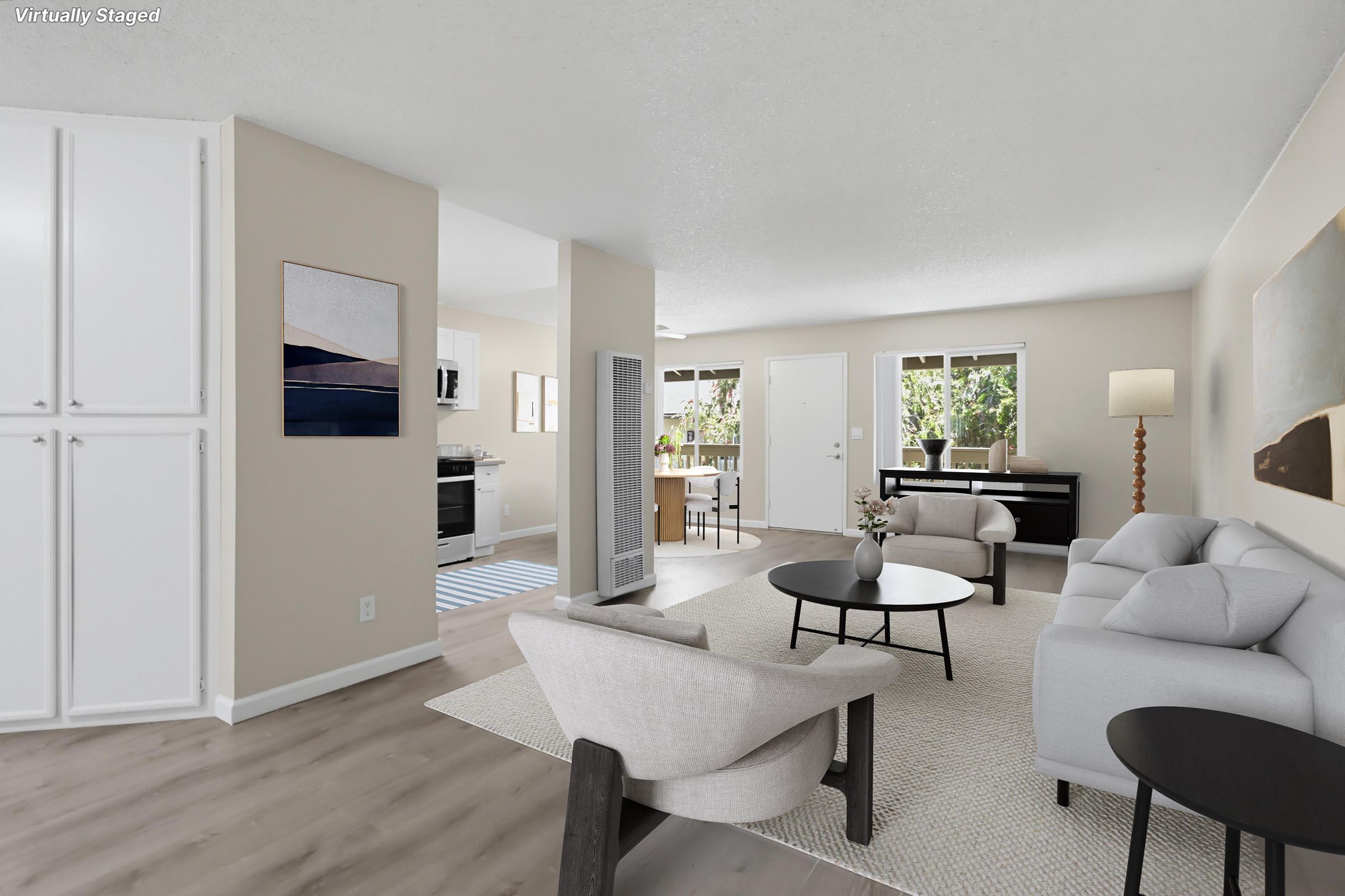 A modern living room featuring light-colored walls and a neutral rug. It includes a sofa, a lounge chair, a round coffee table, and a floor lamp. There’s a piano and a dining area visible in the background, along with large windows allowing natural light to enter the space.