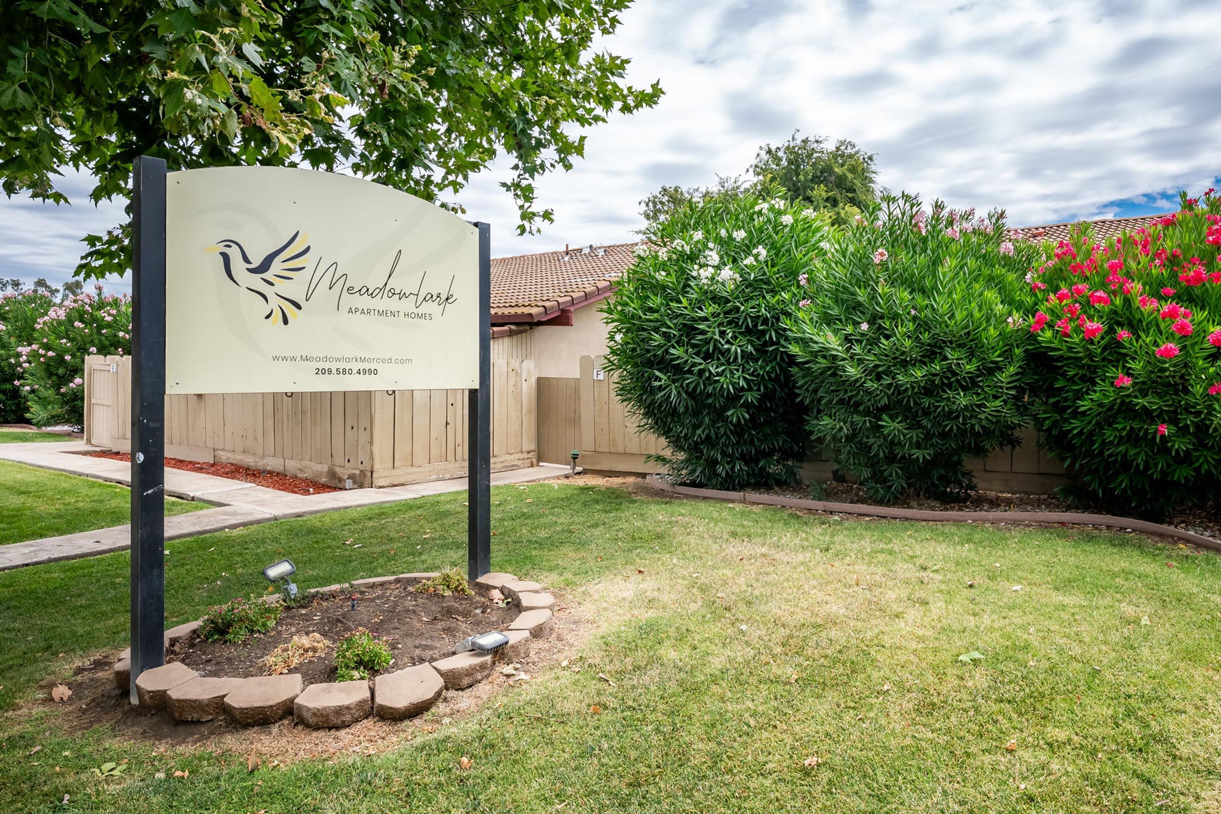 Sign for Meadowlark Apartment Homes, featuring a stylized bird logo. The sign stands in front of a well-maintained landscape with greenery and flowers. There are pathways and a fence in the background, contributing to the welcoming environment of the apartment complex. Cloudy sky overhead.