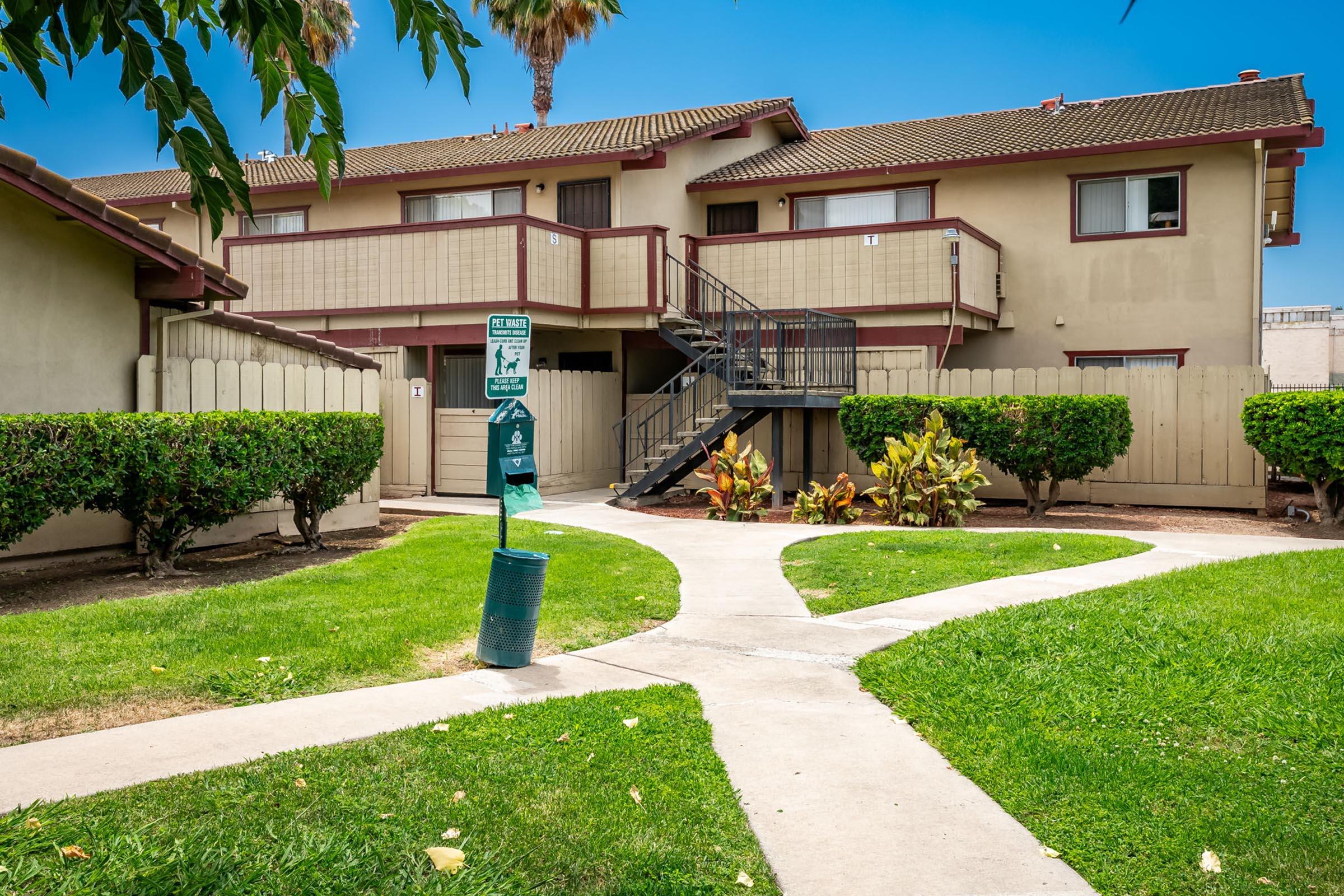 A two-story apartment building with a staircase on the side, surrounded by well-kept green lawns and shrubs. A pathway divides the lawn leading to different areas, with a trash bin visible near the center. Palm trees and a clear blue sky add to the pleasant outdoor environment.