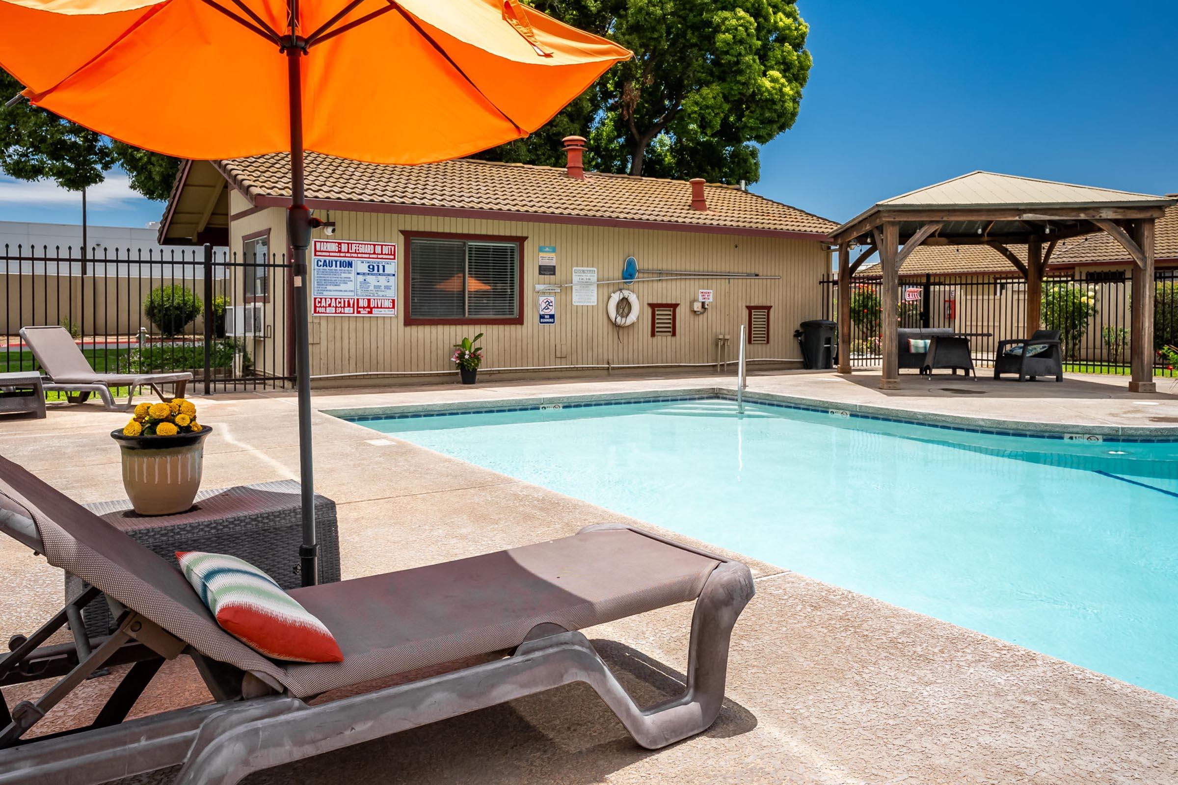 A sunny pool area featuring a clear blue swimming pool with lounge chairs and an orange umbrella. In the background, there is a poolside building with signs and a gazebo. Lush trees and a well-maintained lawn complete the relaxing outdoor atmosphere.