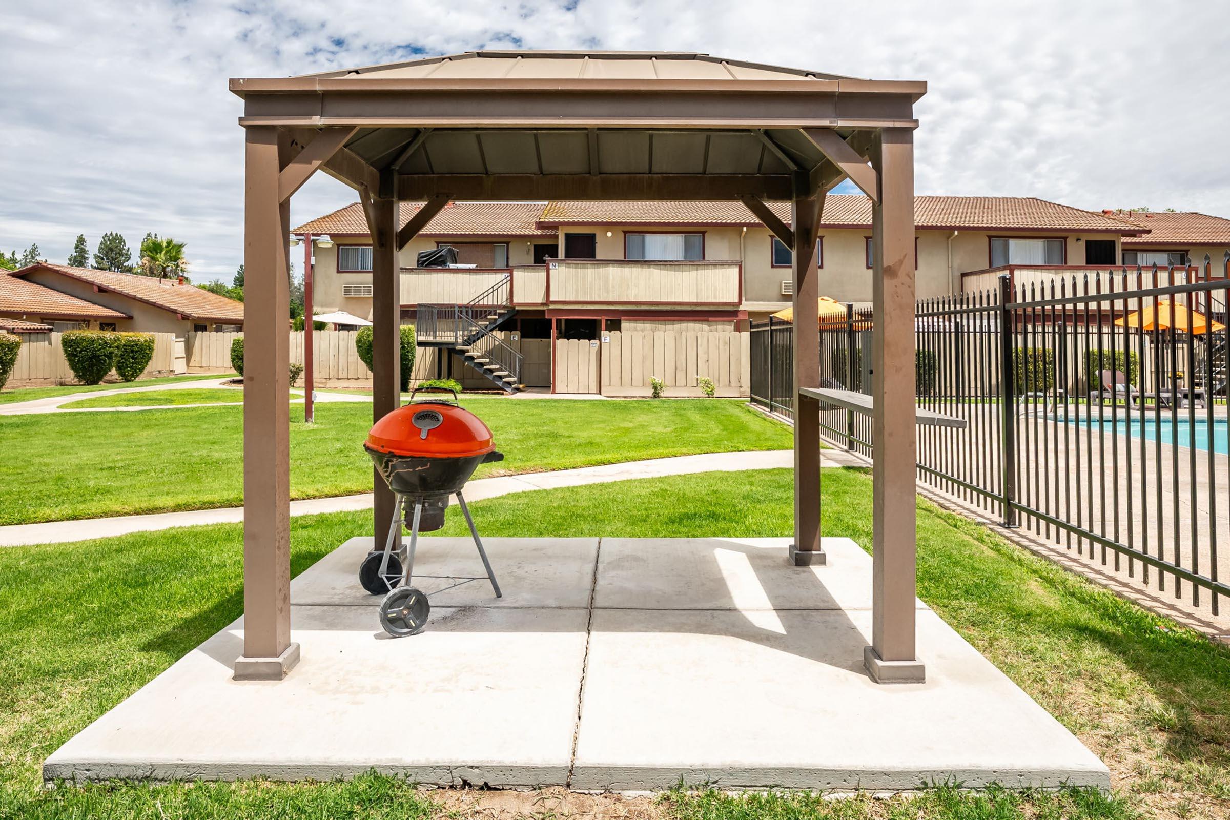 A gazebo with a grill inside, situated on a concrete slab in a green lawn area. In the background, there are two-story residential buildings and a fenced swimming pool area. The sky is partly cloudy, offering a pleasant outdoor environment for cooking and relaxation.