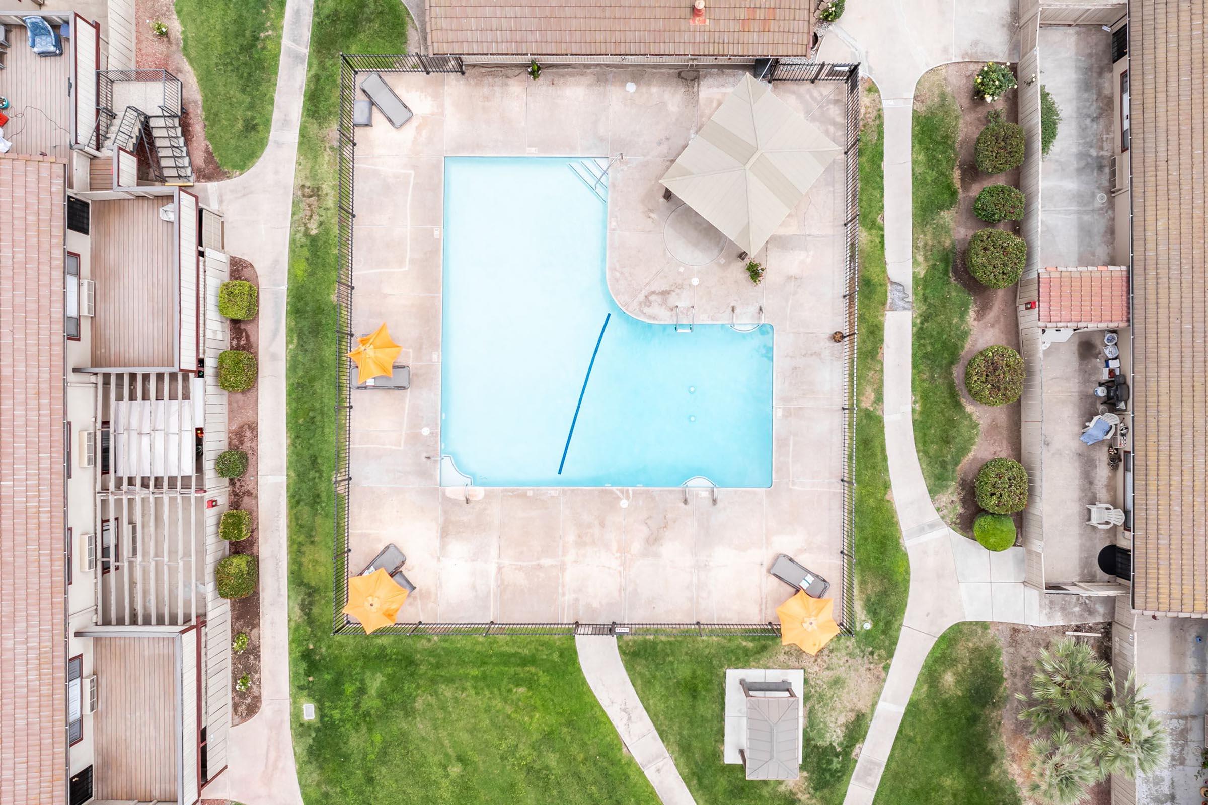 Aerial view of a residential pool area featuring a rectangular swimming pool with a clear blue surface. Surrounding the pool are lounge chairs and yellow umbrellas. Neatly manicured lawns and pathways lead to nearby apartment buildings, creating a community vibe.