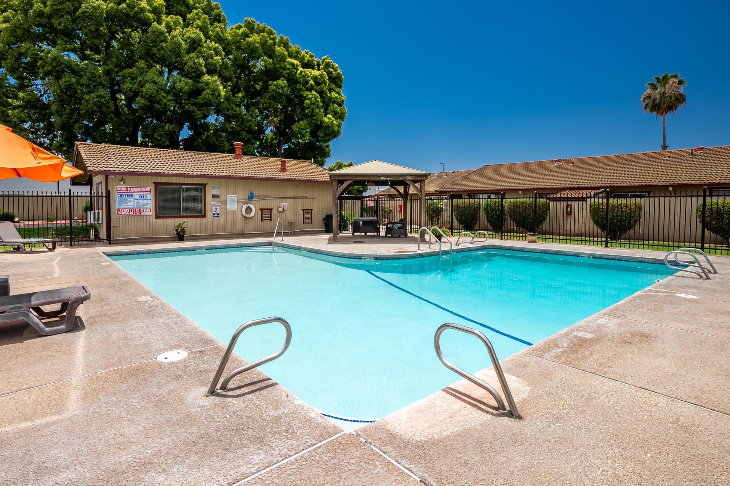 A clear swimming pool with a shallow end and ladders on a sunny day. Surrounding the pool are lounge chairs and umbrellas, with a fenced area and several trees in the background, creating a relaxing outdoor atmosphere. A small canopy with seating is also visible nearby.