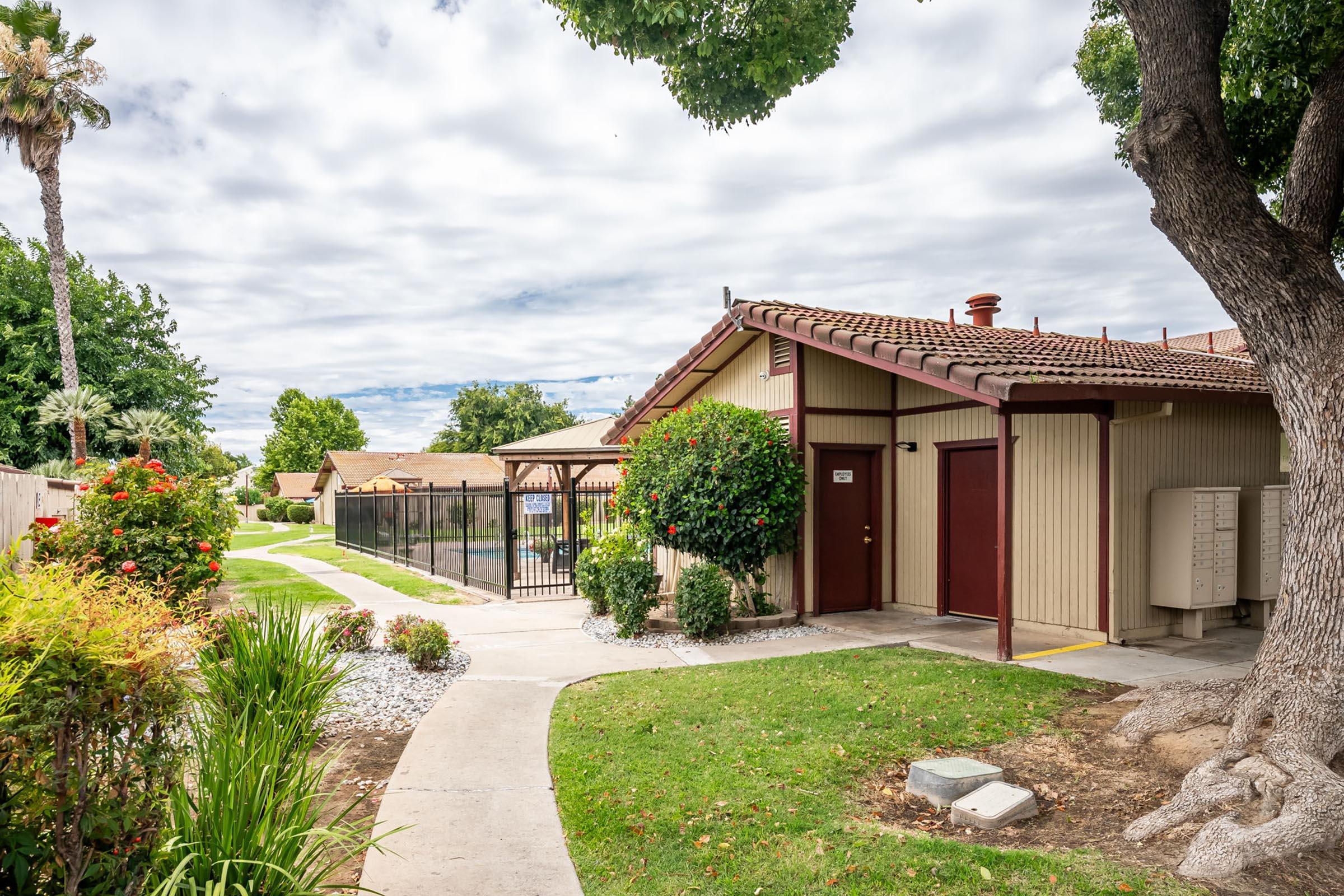 A pathway lined with green grass and shrubs leads to a small building with a reddish-brown roof and beige walls. The building has two doors, and a fenced area is visible in the background, surrounded by trees and vegetation. The sky is partly cloudy, adding to the serene atmosphere.