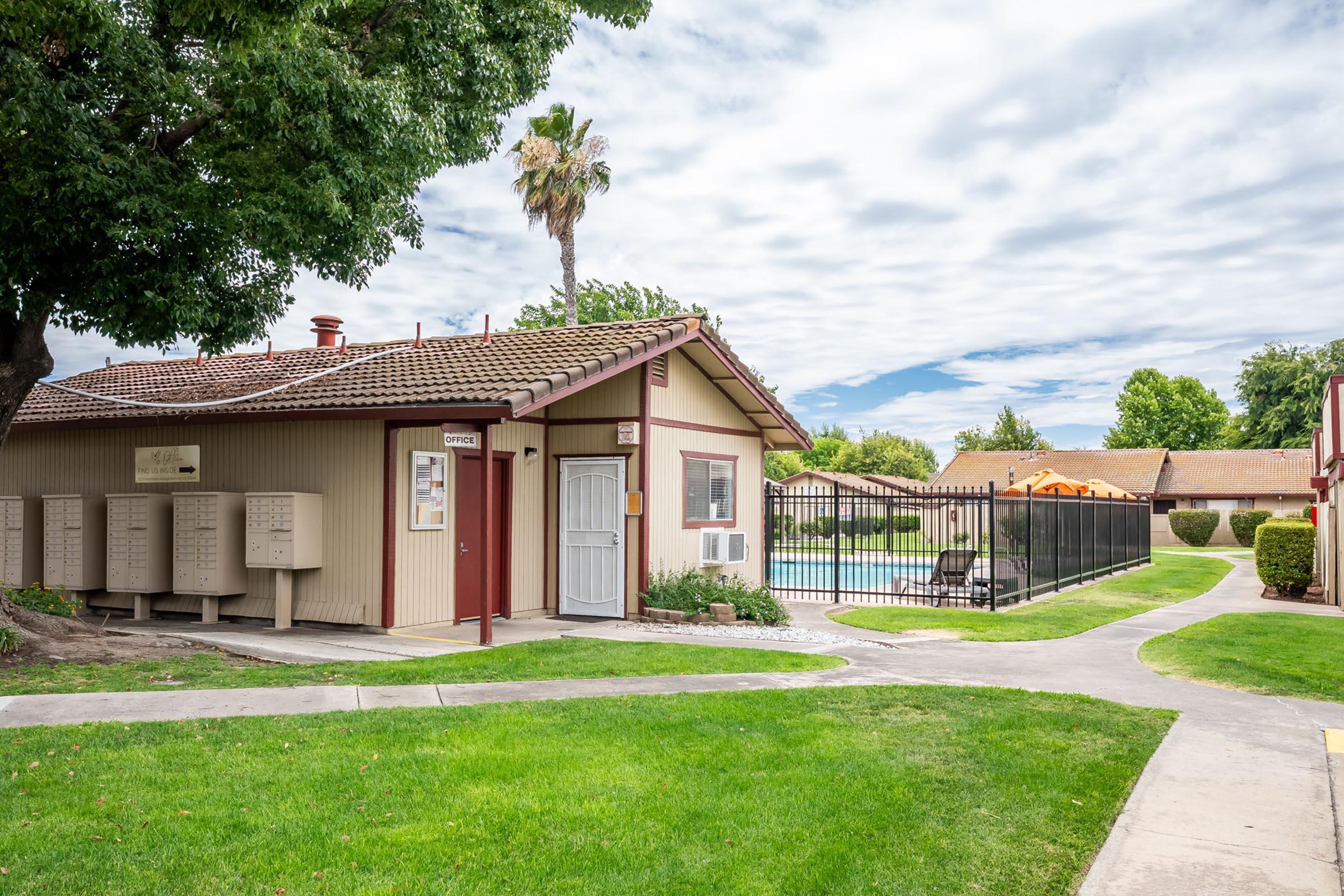 A residential area featuring a small building with mailboxes, a swimming pool enclosed by a black fence, and landscaped green spaces. Palm trees and a cloudy sky are visible, creating a peaceful atmosphere in the neighborhood. A path leads through the lawn towards the pool area.
