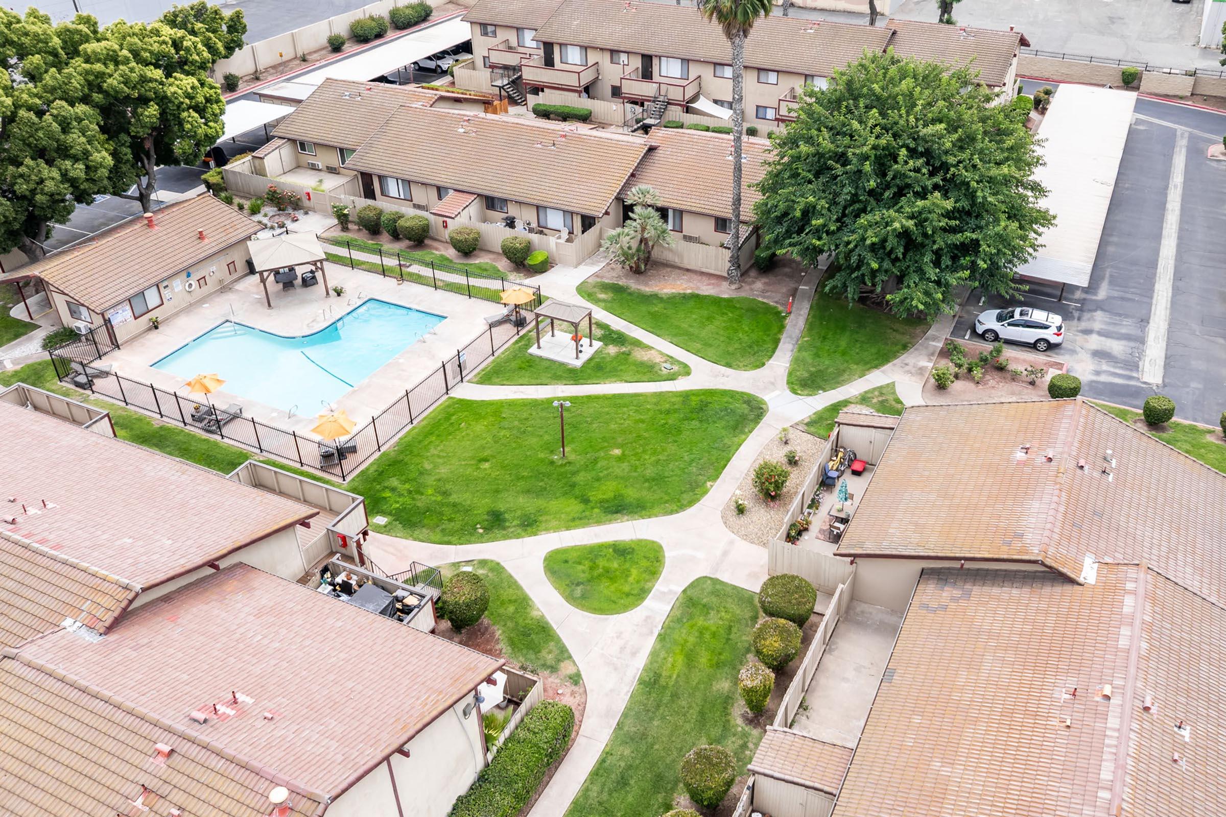 An aerial view of a residential complex featuring a central pool surrounded by neatly landscaped green lawns and pathways. Several buildings with brown roofs are visible, along with outdoor furniture near the pool area. The surroundings include trees and parking spaces.