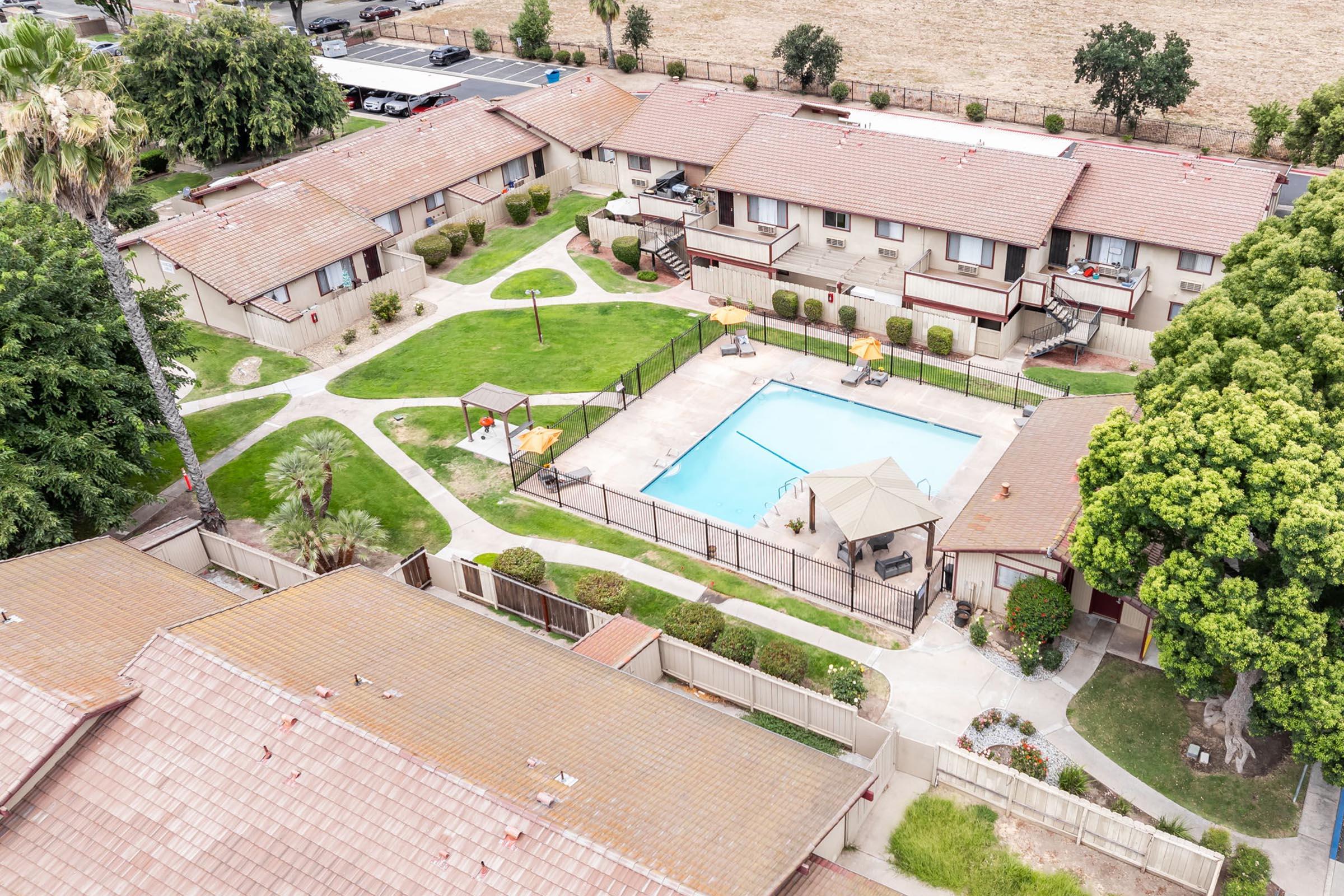 Aerial view of a residential complex featuring several two-story buildings surrounding a central area with a rectangular swimming pool. The pool is enclosed by a fence and has lounge chairs. Lush green lawns and trees add to the landscape, creating a relaxing outdoor space.