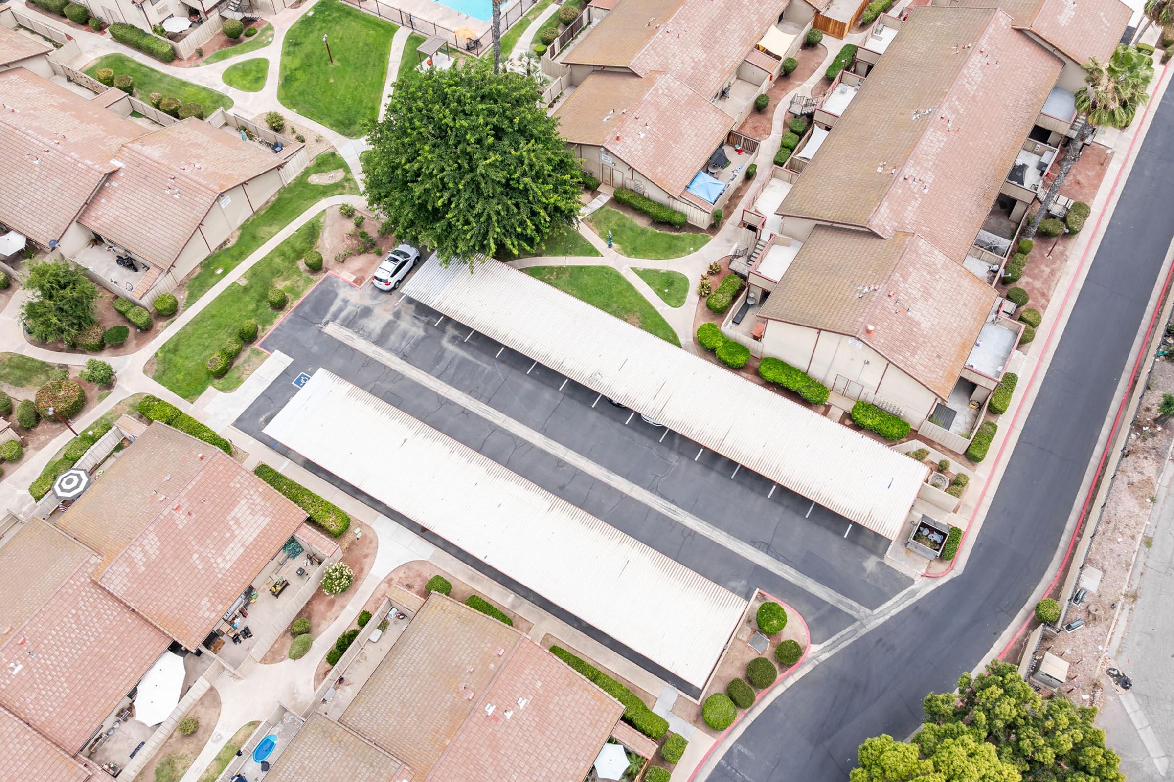 Aerial view of a residential area featuring several one-story apartment buildings with flat roofs. Pathways and greenery are visible, along with a parking lot that has covered spaces. A few trees are scattered throughout the landscape, and a clear blue sky adds to the overall scenic appearance.