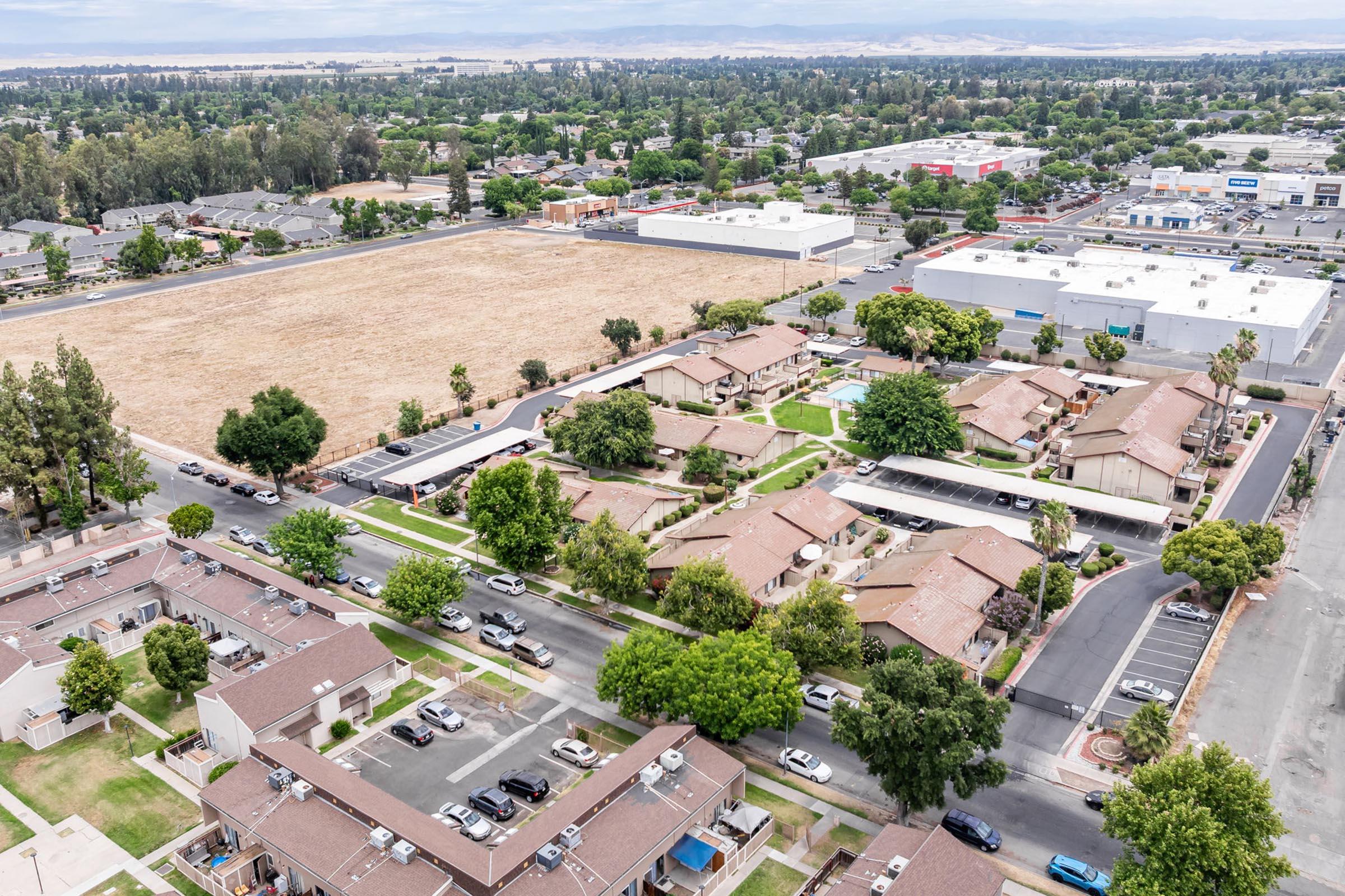 Aerial view of a suburban neighborhood featuring single-story apartment buildings surrounded by trees and parking areas. In the background, there's an empty field and a commercial area with large storefronts. The landscape includes greenery and residential units, showcasing a mix of developed and undeveloped land.