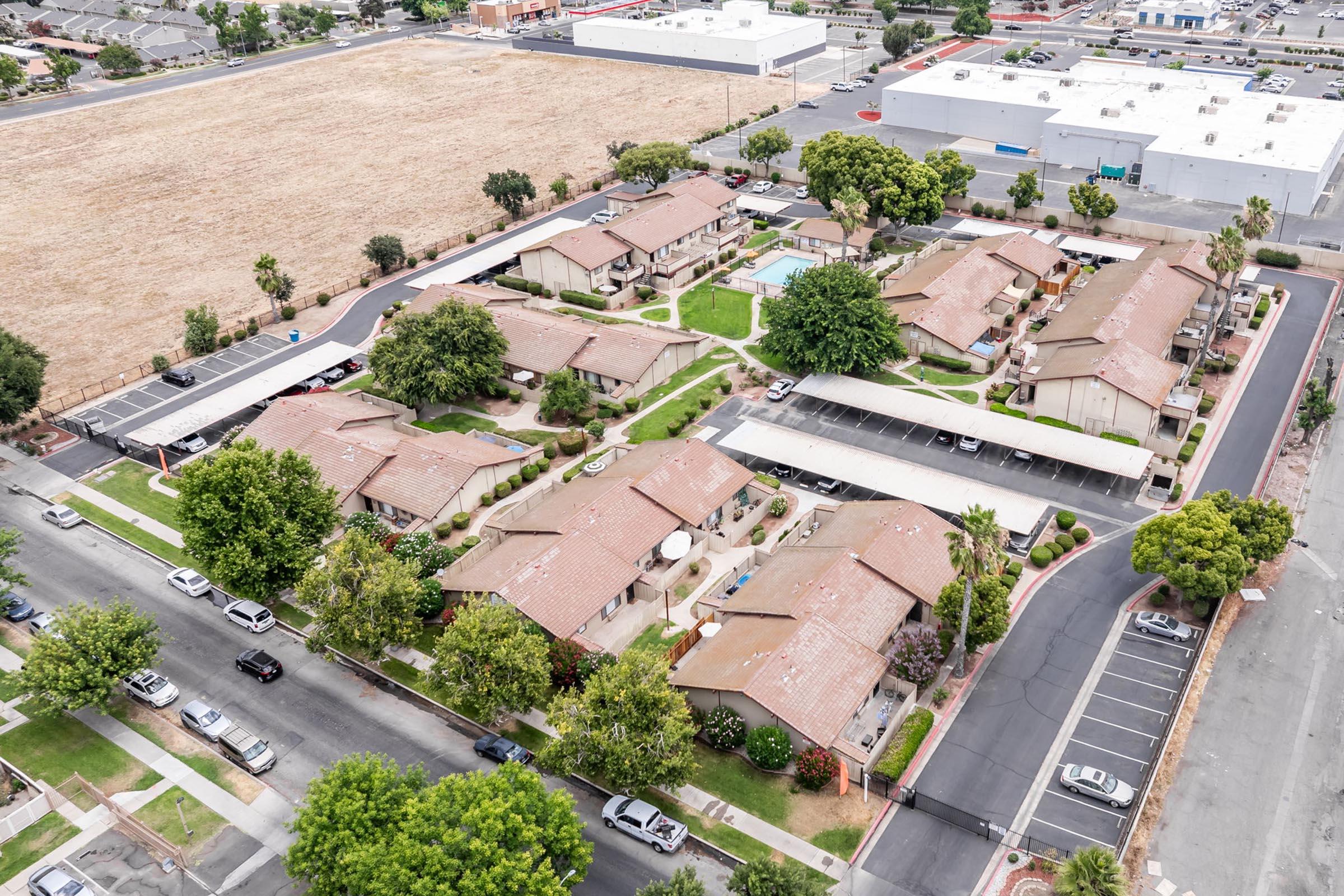 Aerial view of a residential complex featuring multiple single-story buildings with red roofs, surrounded by green landscaping, trees, and a parking area. In the background, a large commercial building and an open field are visible. The scene captures a suburban environment with well-maintained grounds.