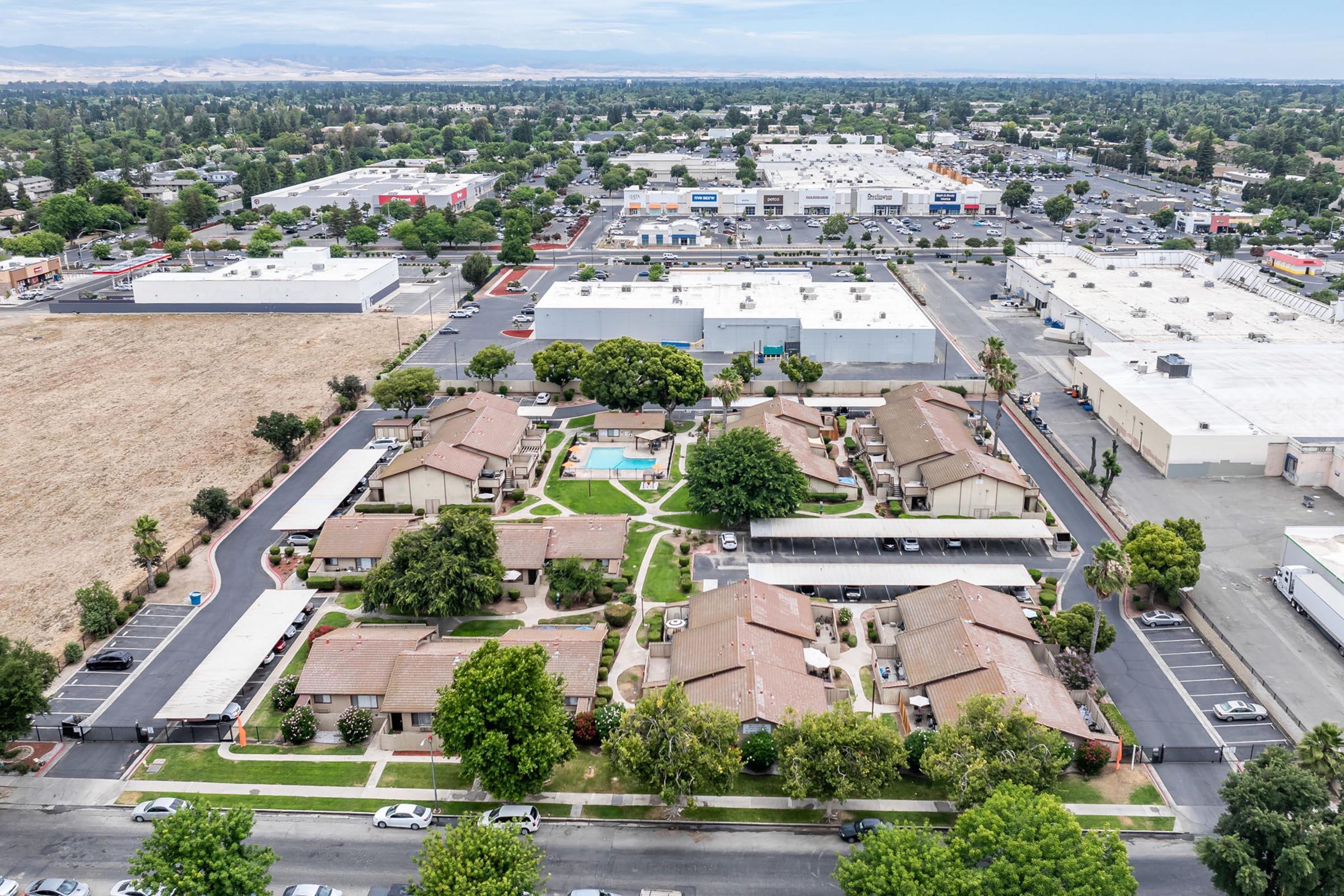 An aerial view of a residential complex featuring several single-story buildings arranged around a central pool area, surrounded by landscaping. In the background, a commercial area with large parking lots and retail stores is visible, along with distant mountains.