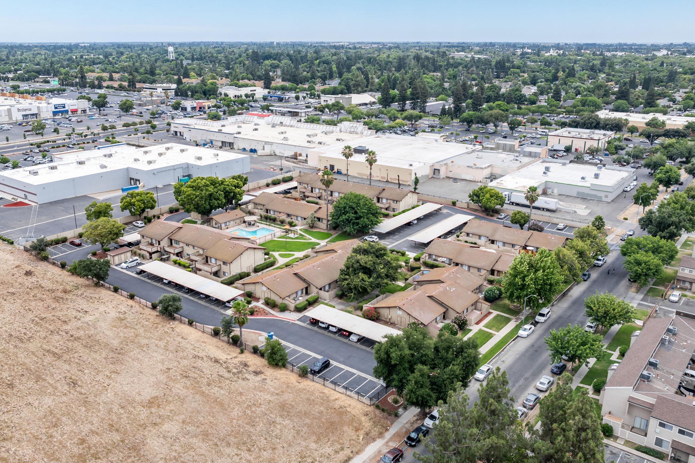 Aerial view of a residential complex surrounded by green trees and landscaped areas, with single-story buildings arranged around a central courtyard. In the background, there are commercial buildings and parking lots, indicative of a suburban setting. The area features a mix of housing and commercial development.