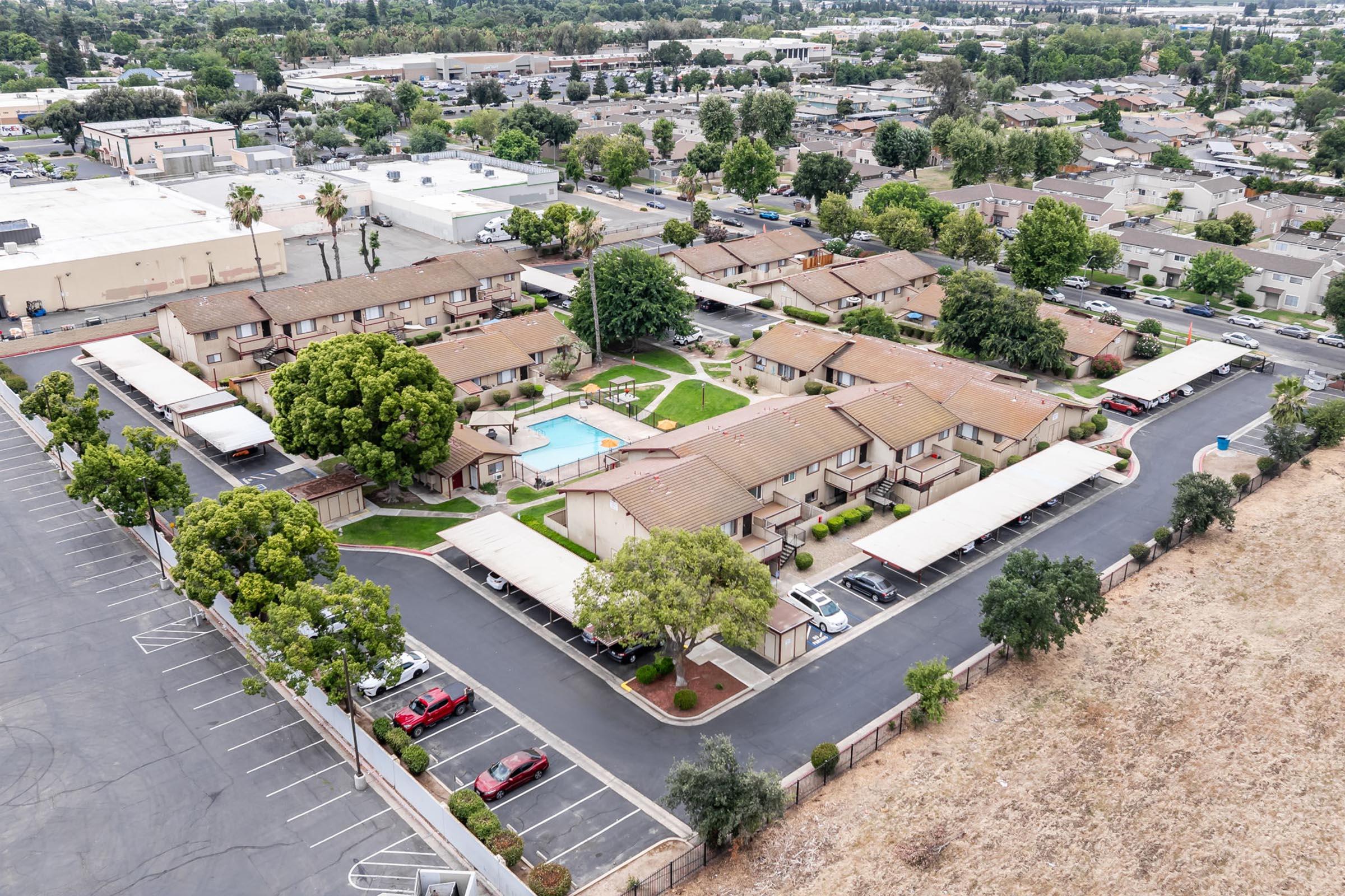 Aerial view of a residential complex featuring multiple single-story buildings arranged around a central swimming pool. The surrounding area includes parking spaces and landscaped trees. In the background, there are additional commercial buildings and a suburban neighborhood.