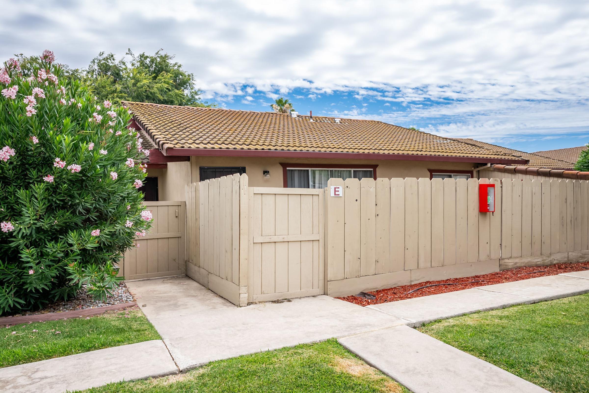 A single-story house with a brown tiled roof, surrounded by a wooden fence. The fence is partially open, revealing a red mailbox near the entrance. Lush green shrubs with pink flowers are visible on the left, and a well-maintained lawn and sidewalk lead up to the house under a partly cloudy sky.