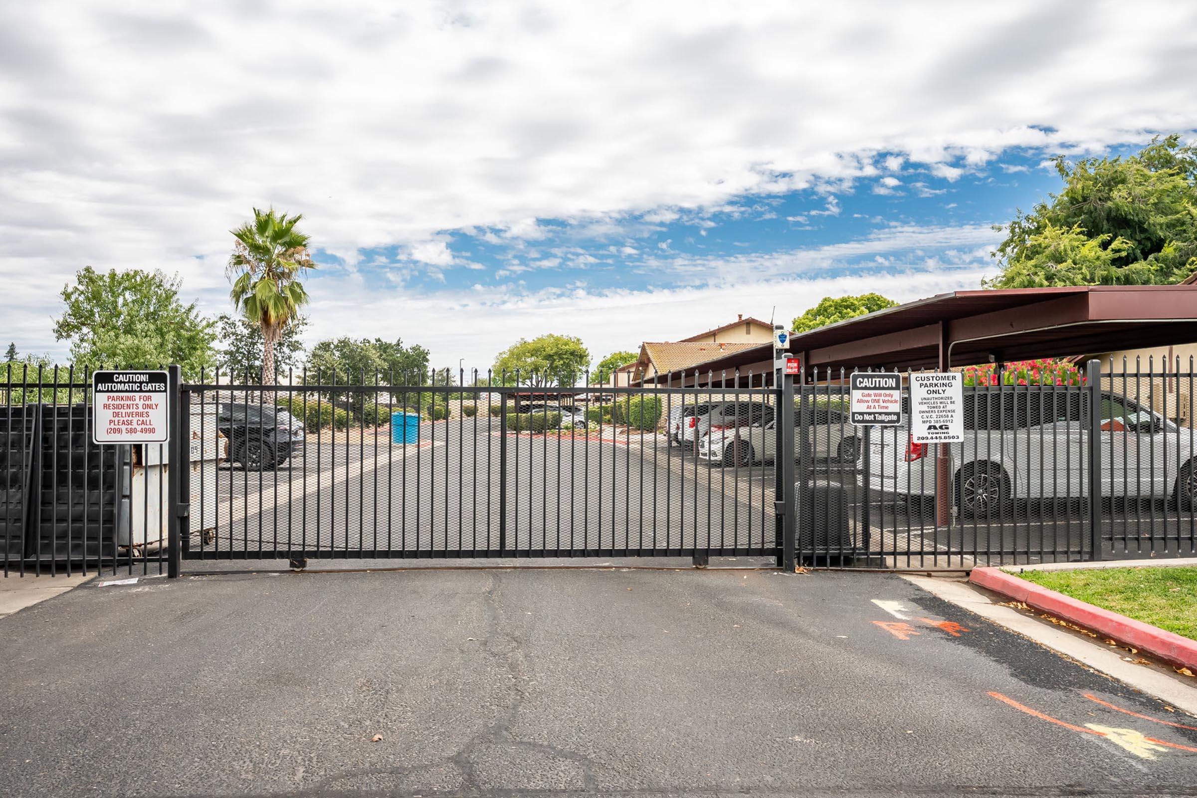 A gated entrance to a parking area, featuring a black metal gate. The scene includes several parked cars, palm trees in the background, and a cloudy sky. Warning signs are posted on the gate, indicating restricted access. The pathway is paved and bordered with red curbing.