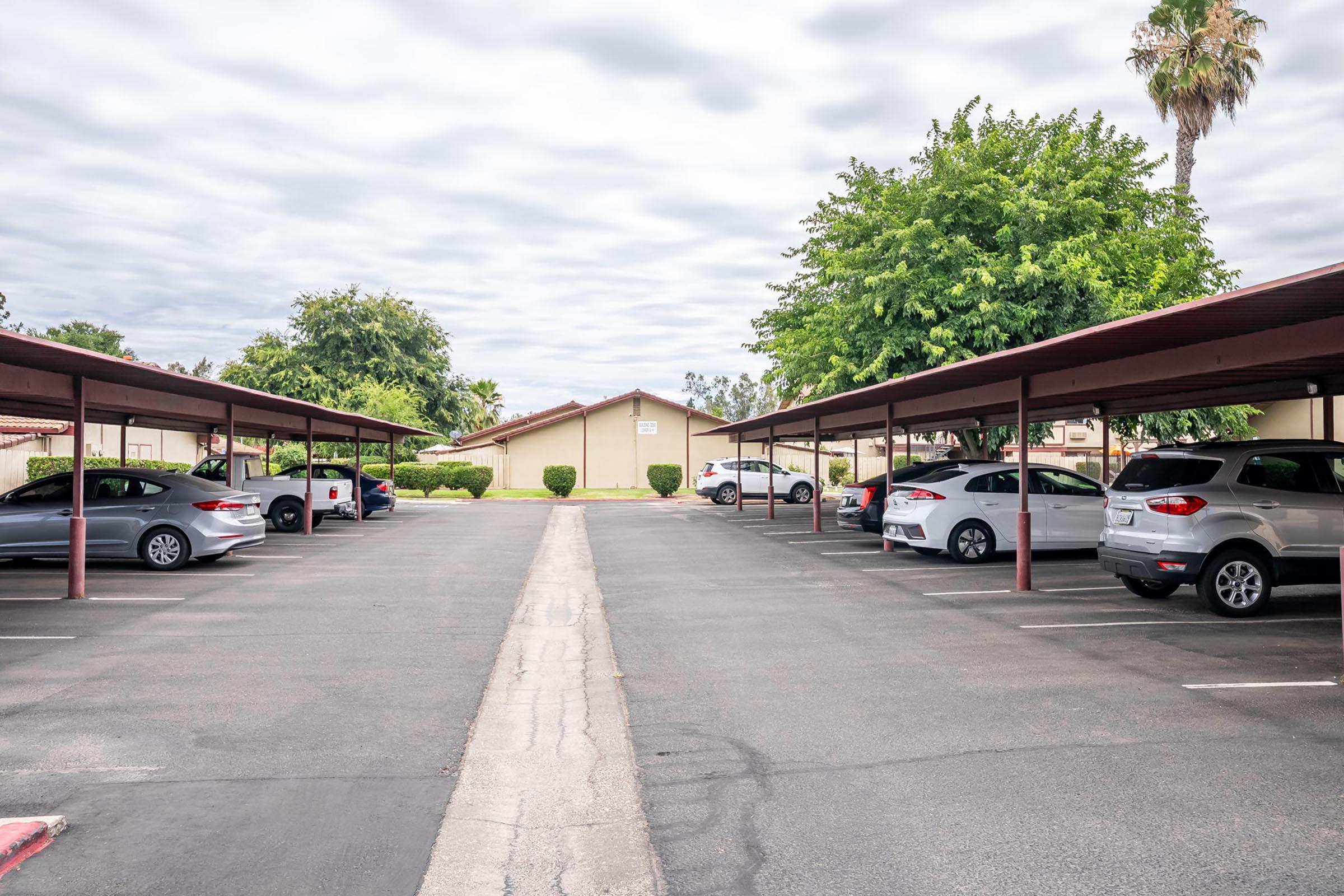 A wide view of a parking lot with covered parking spaces. Several cars are parked under the covers, and there are trees and a building in the background. The sky is overcast, creating a subdued light over the scene.