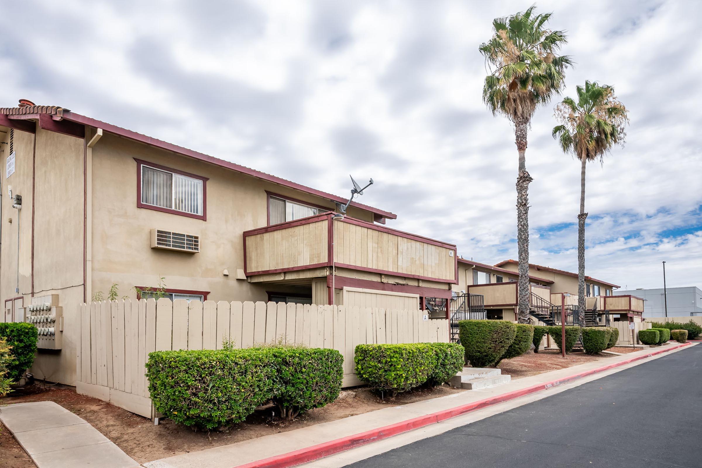 A low-rise apartment complex with a beige exterior, featuring balconies and windows. Lush green shrubs line the walkway and a wooden fence borders the property. Two tall palm trees stand in the foreground under a partly cloudy sky.