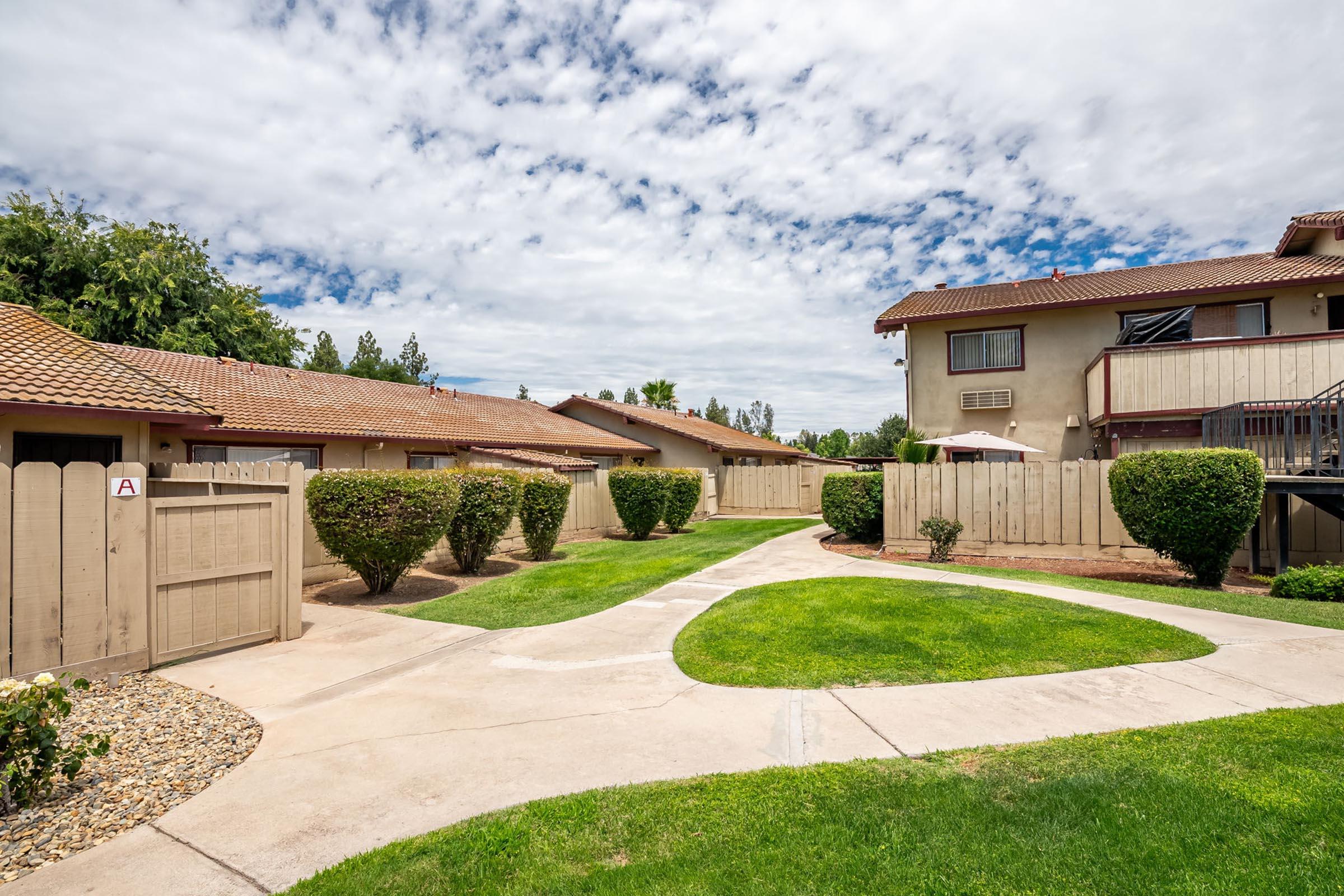 A landscaped pathway winds between low wooden fences and shrubs in a residential area. Two-story buildings with balconies are visible in the background under a partly cloudy sky. Green grass surrounds the walkway, creating a calm and inviting atmosphere.