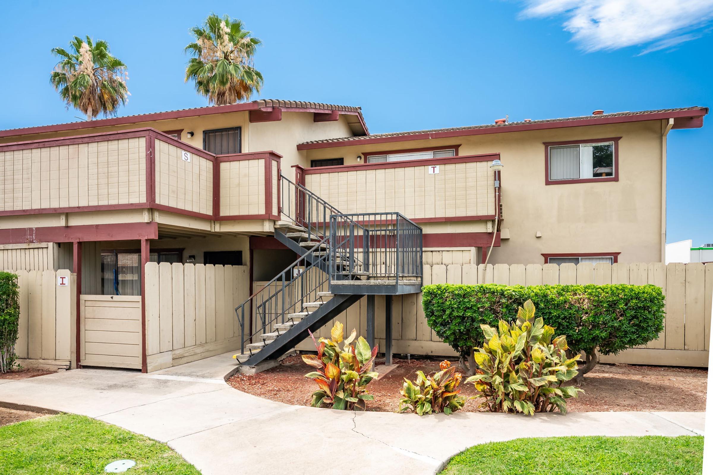 An apartment building with a two-story structure featuring a staircase on the side. The exterior is tan with red accents. There are palm trees in the background and a landscaped area with shrubs and plants in the foreground. The sky is clear and blue.