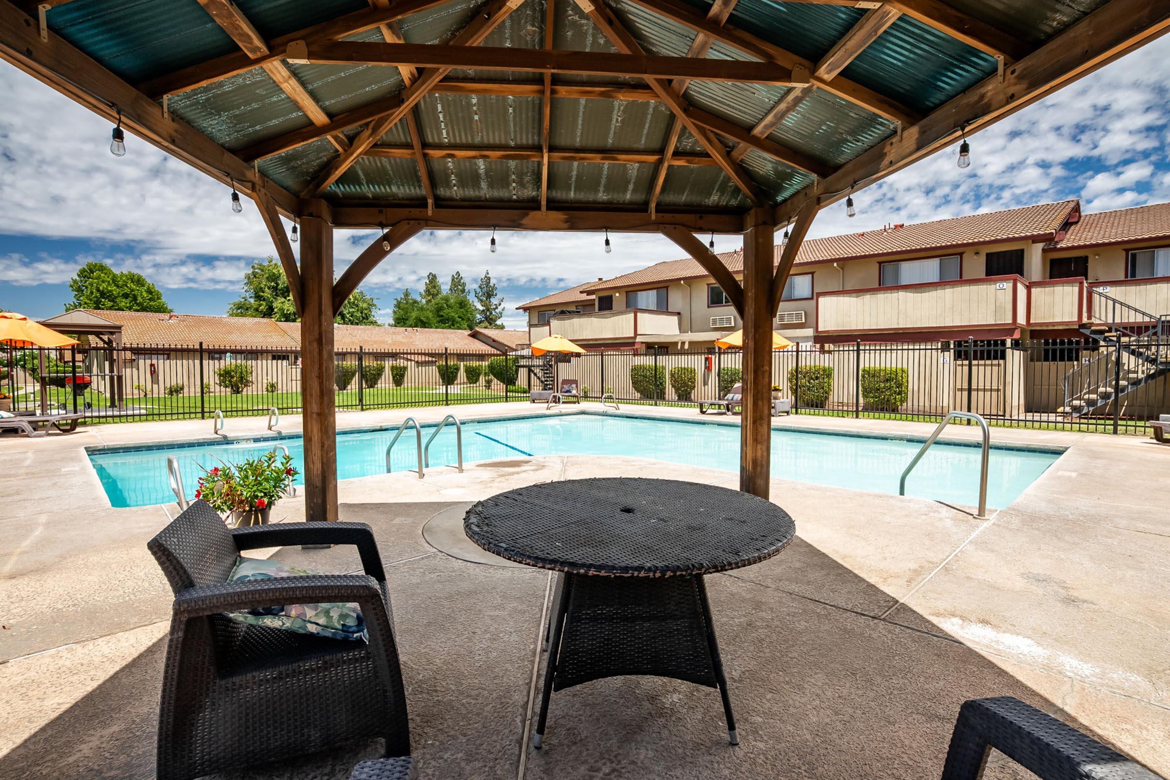 A shaded gazebo with a round table and chairs overlooks a sparkling swimming pool. Surrounding the pool are lounge chairs and umbrellas, with a backdrop of multi-story apartment buildings and a partly cloudy blue sky. The scene conveys a relaxing outdoor atmosphere.
