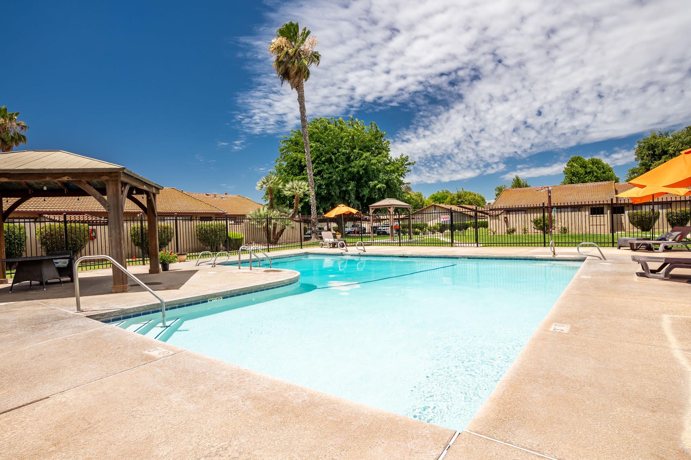 A bright, sunny pool area featuring a clear blue swimming pool surrounded by lounge chairs and umbrellas. In the background, there are green trees and residential buildings under a partly cloudy sky, creating a relaxing atmosphere for leisure and recreation.