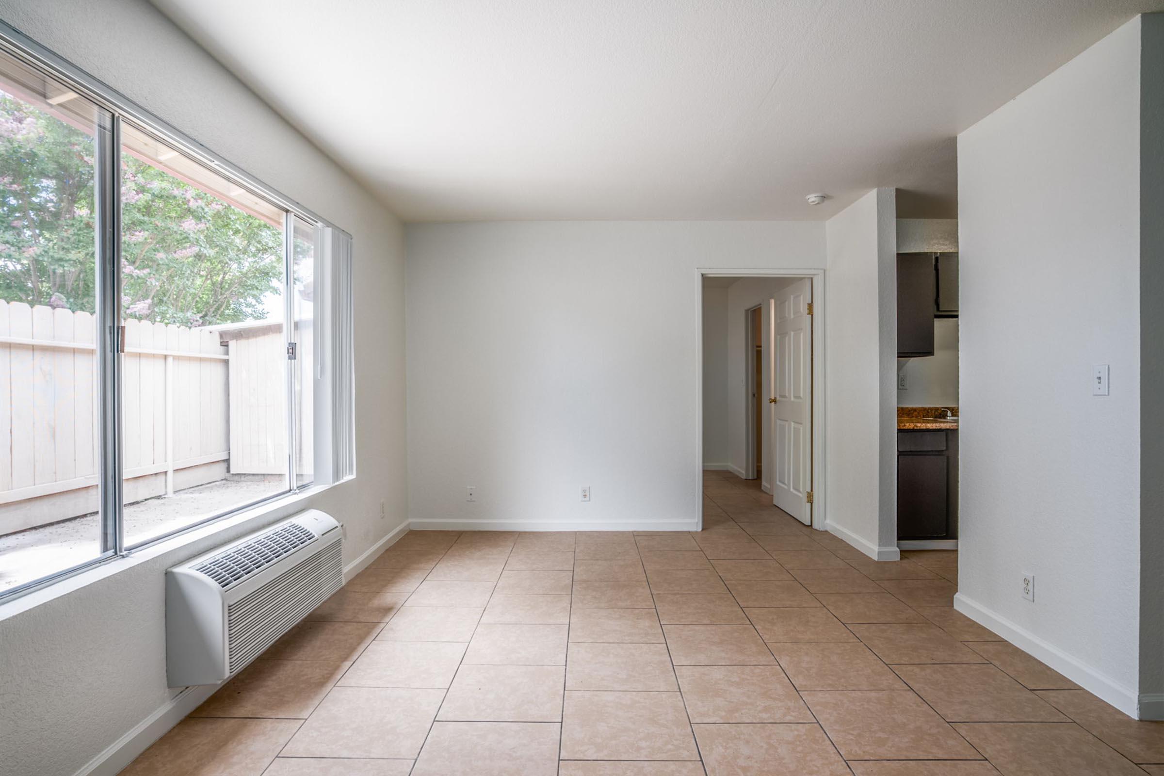 Bright, empty living room featuring large windows, light-colored walls, tiled flooring, and an air conditioning unit. A doorway leads to a hallway and kitchen area. Natural light fills the space, which has a minimalist and open feel. The room is ready for furniture and personalization.