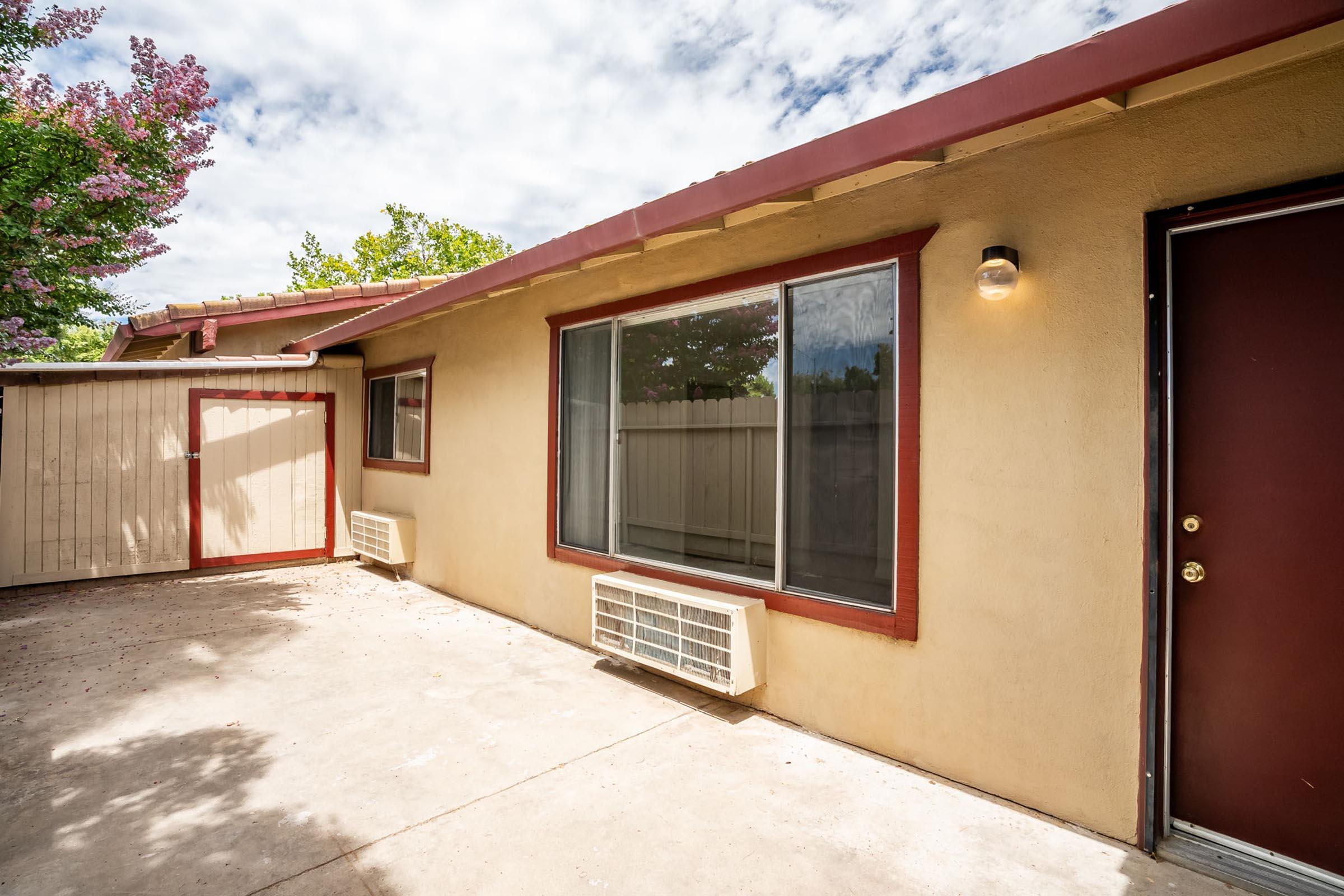 A view of a house exterior featuring a beige wall, a large window with red trim, and an air conditioning unit below it. The entrance door is dark brown. To the side, there is a small shed or storage space. The area is partly shaded by trees and a cloudy sky can be seen above.
