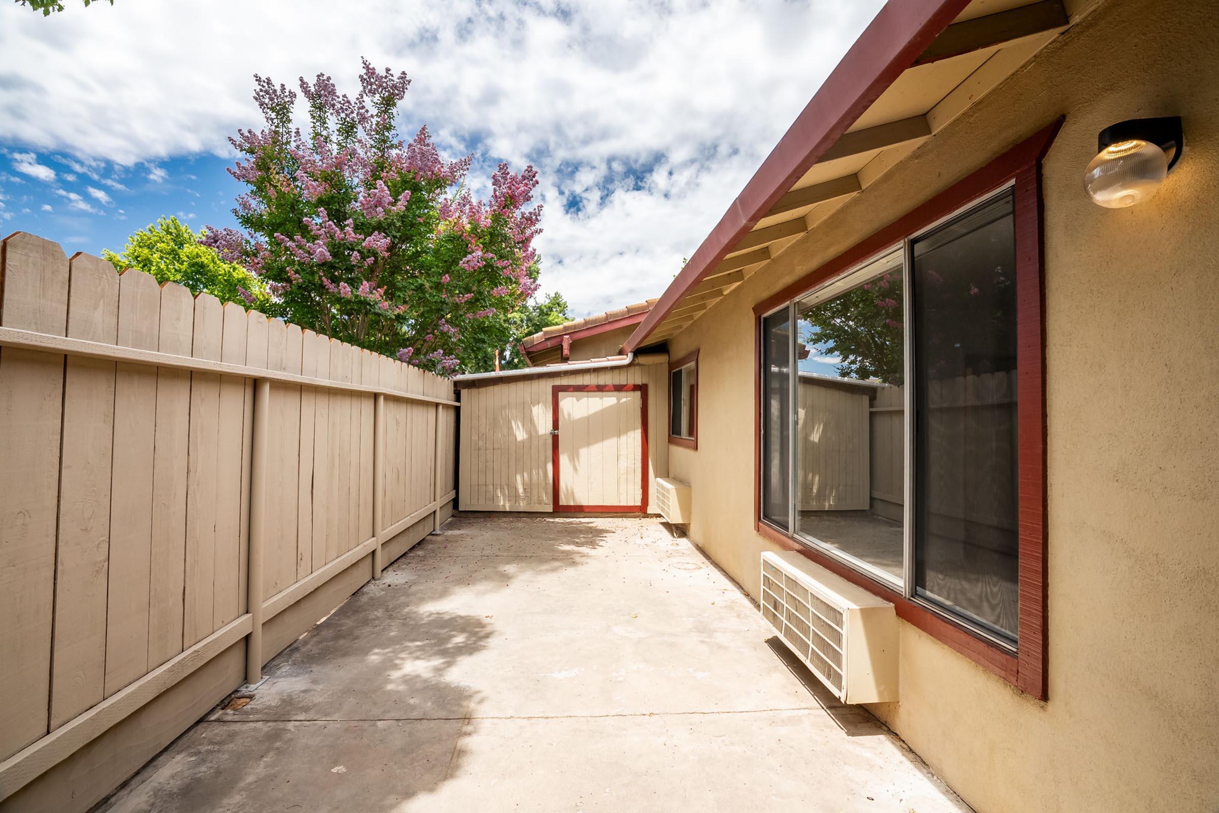 A small outdoor patio area with a concrete floor, surrounded by a wooden fence. There is a flower bush with pink blooms nearby, and a door leading to an enclosed space. The sky is partly cloudy, indicating a bright day. Air conditioning unit visible on the wall.