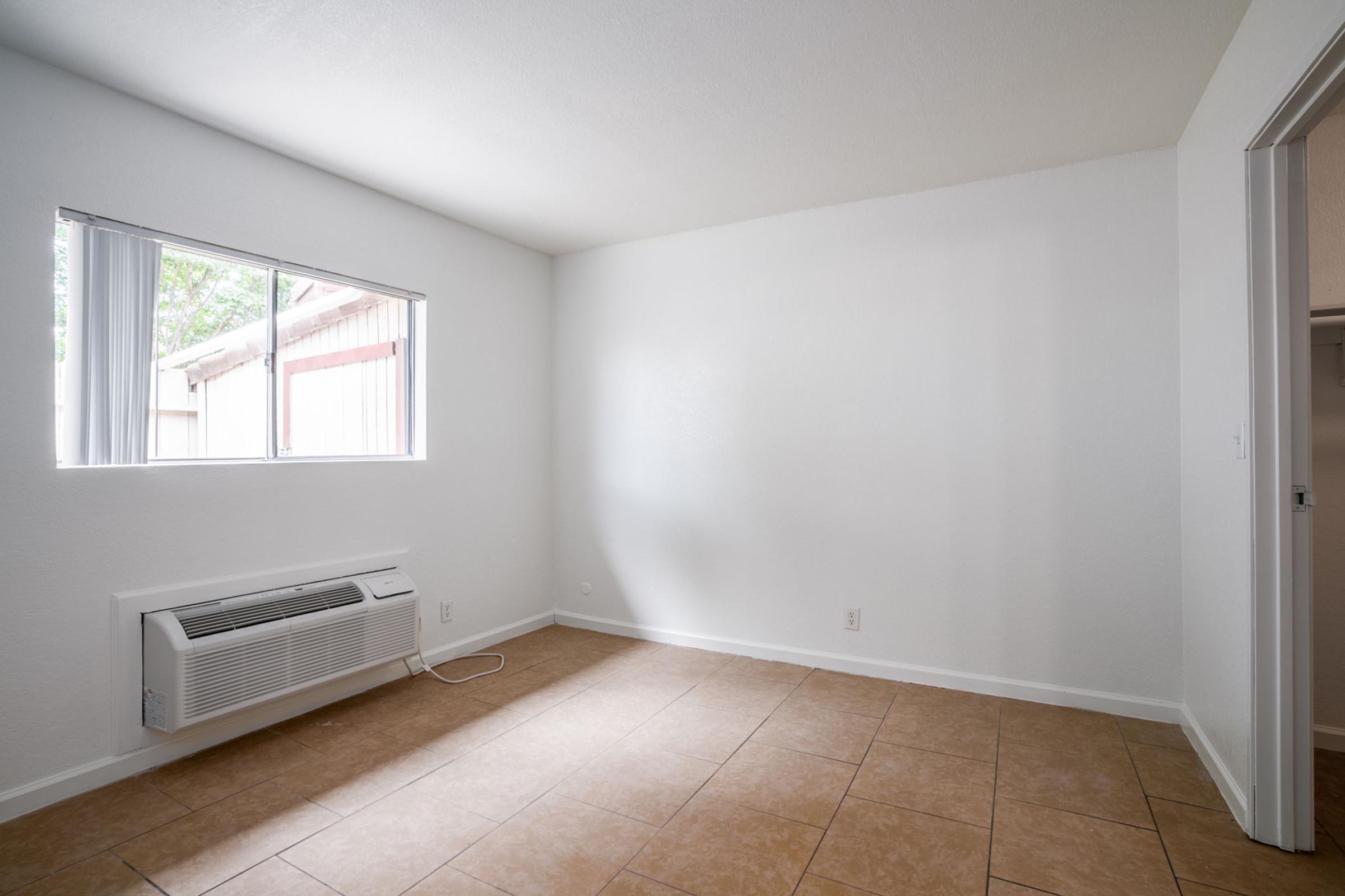 A small, empty room with light-colored walls and tiled flooring. A window allows natural light to enter, while an air conditioning unit is mounted on the wall below the window. The space is uncluttered, with a doorway leading to another area visible in the background.