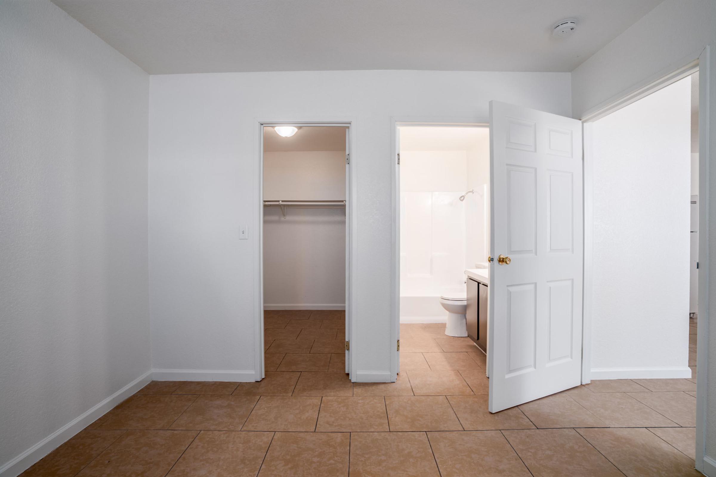 A view of a simple, empty room with tile flooring. Two doors are visible: one leading to a closet and the other to a bathroom. The walls are painted white, and there is a light fixture on the ceiling. The bathroom shows a bathtub and toilet through an open door.