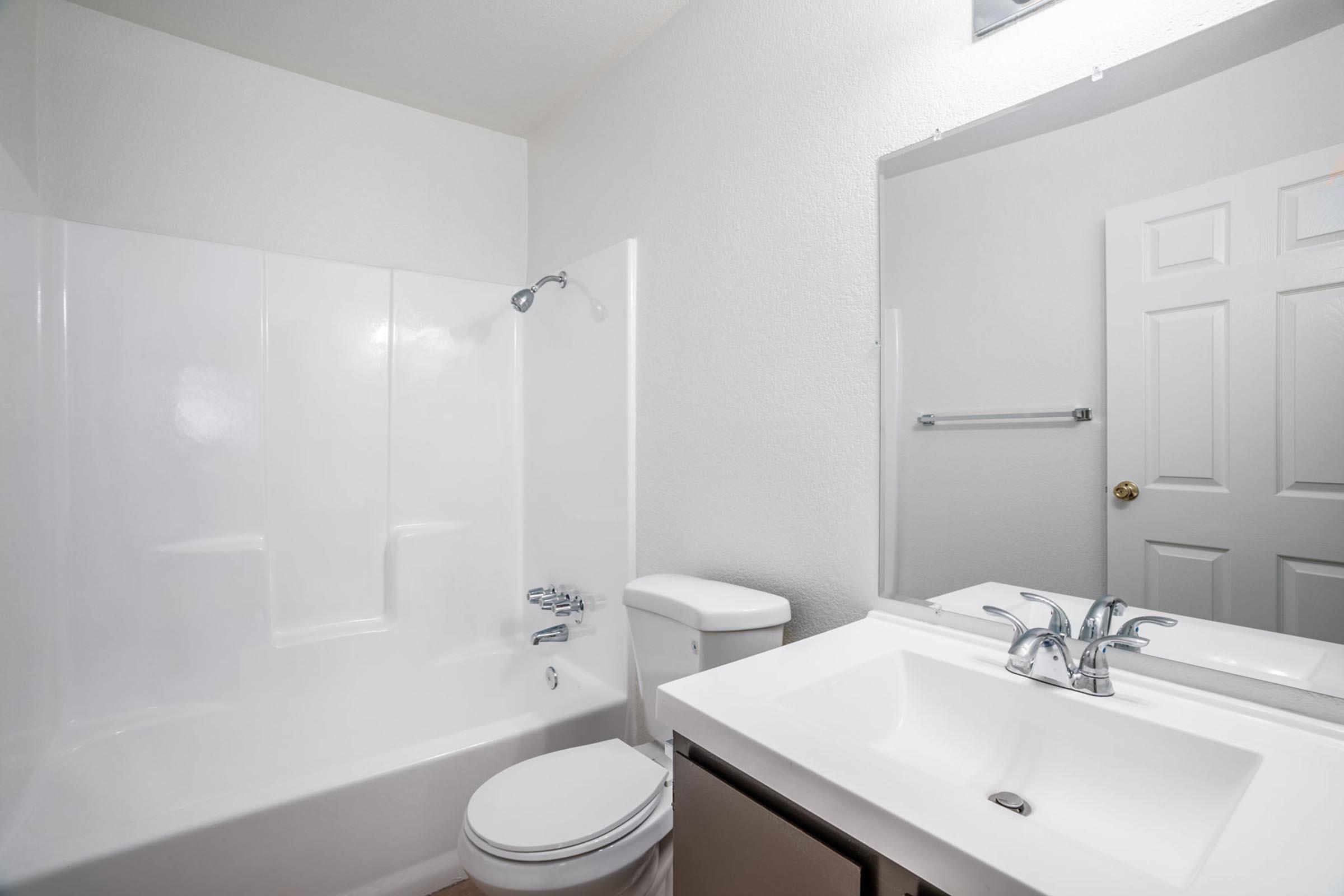 A clean and simple bathroom featuring a white bathtub with a showerhead, a white toilet, and a modern sink with a chrome faucet. The walls are painted light-colored, and there's a mirror above the sink, as well as a door leading to another room, enhancing the overall brightness and spacious feel of the space.