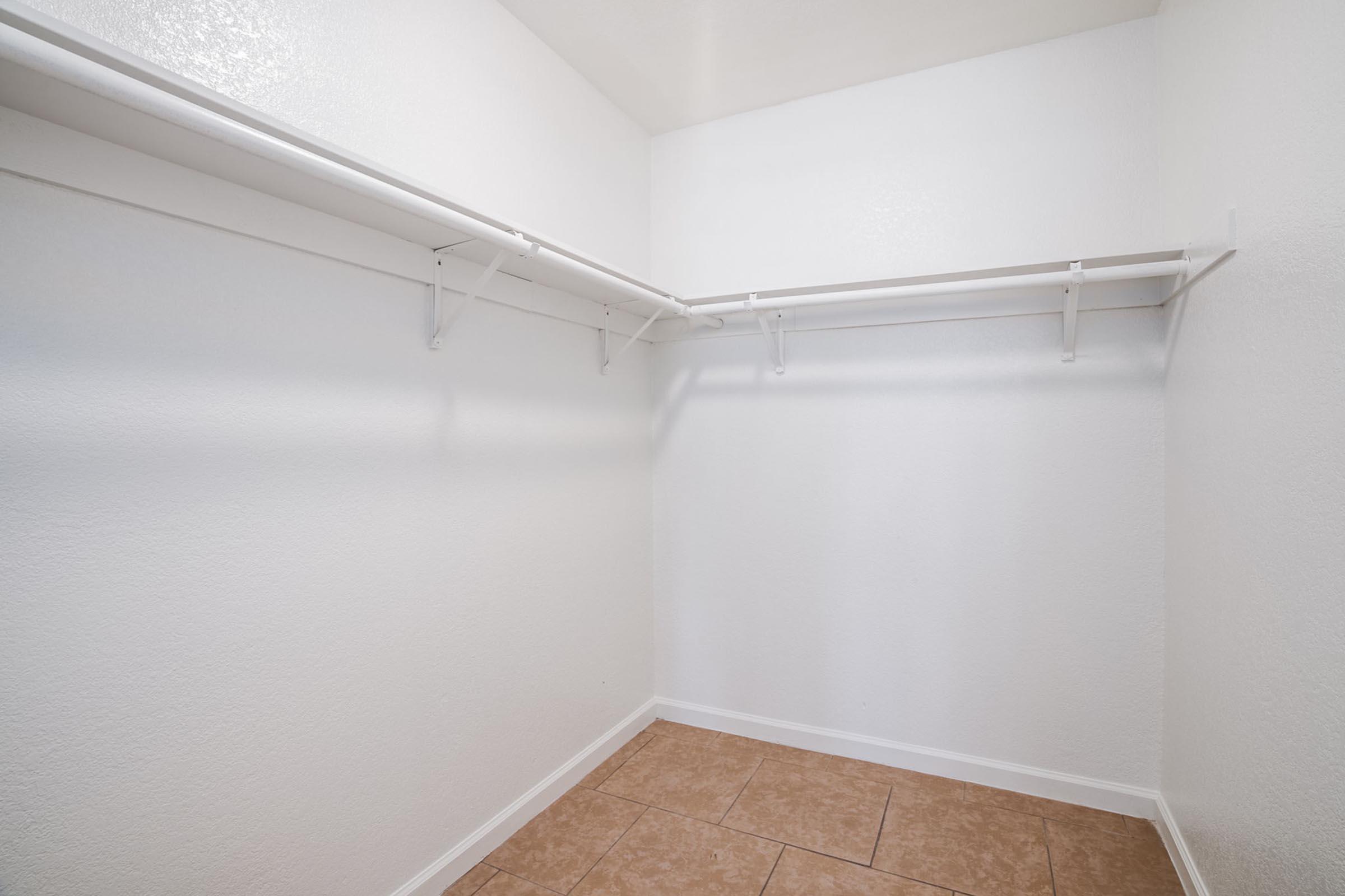 A spacious, empty walk-in closet featuring white walls, light-colored tile flooring, and two white shelving units along the wall. The closet is uncluttered, creating a bright and open atmosphere.