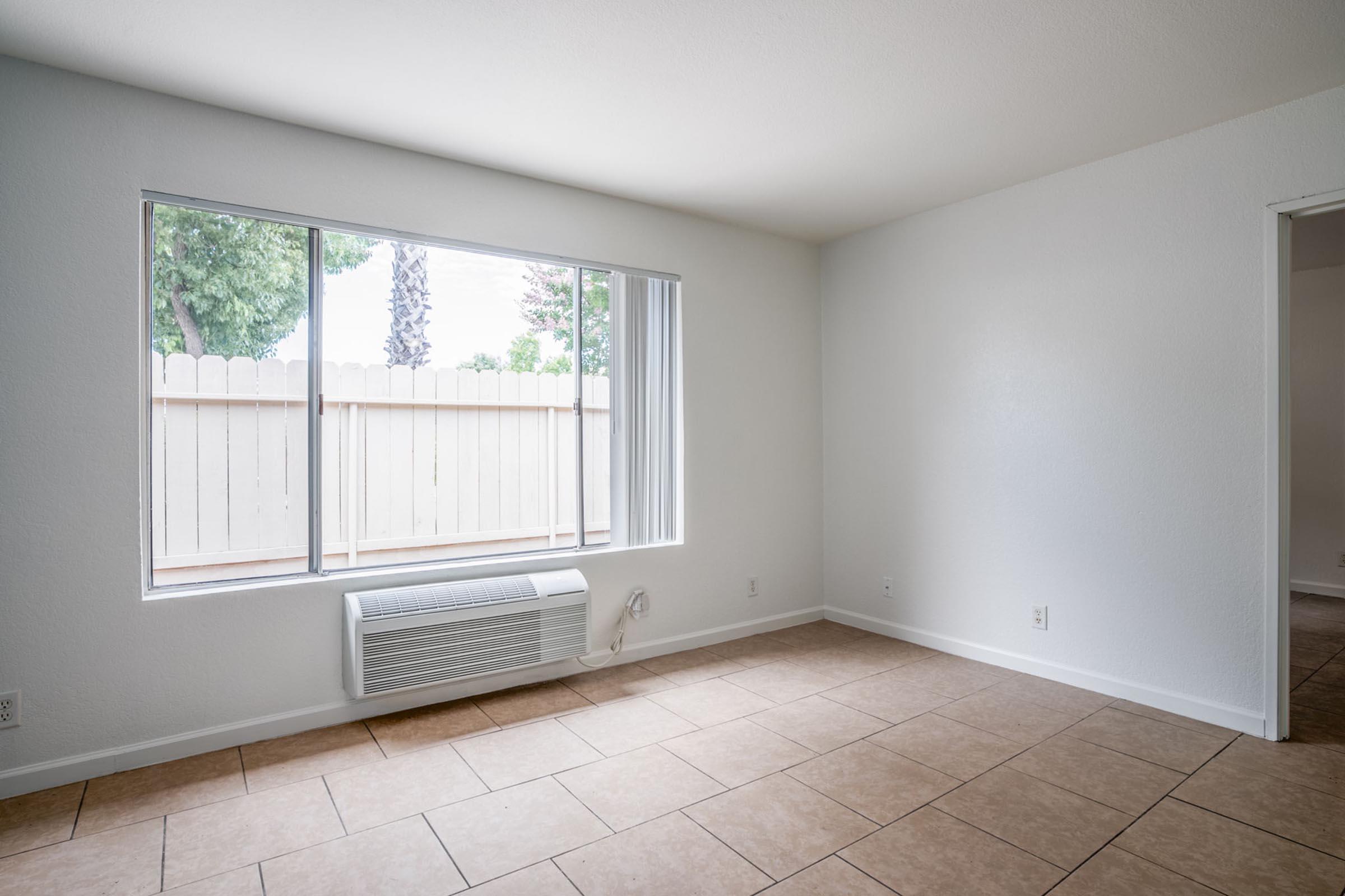 A bright, unfurnished room with light-colored walls and tiled flooring. A large window lets in natural light, facing a fenced outdoor area with trees visible outside. An air conditioning unit is mounted below the window, and there is an open doorway leading to another room or space.