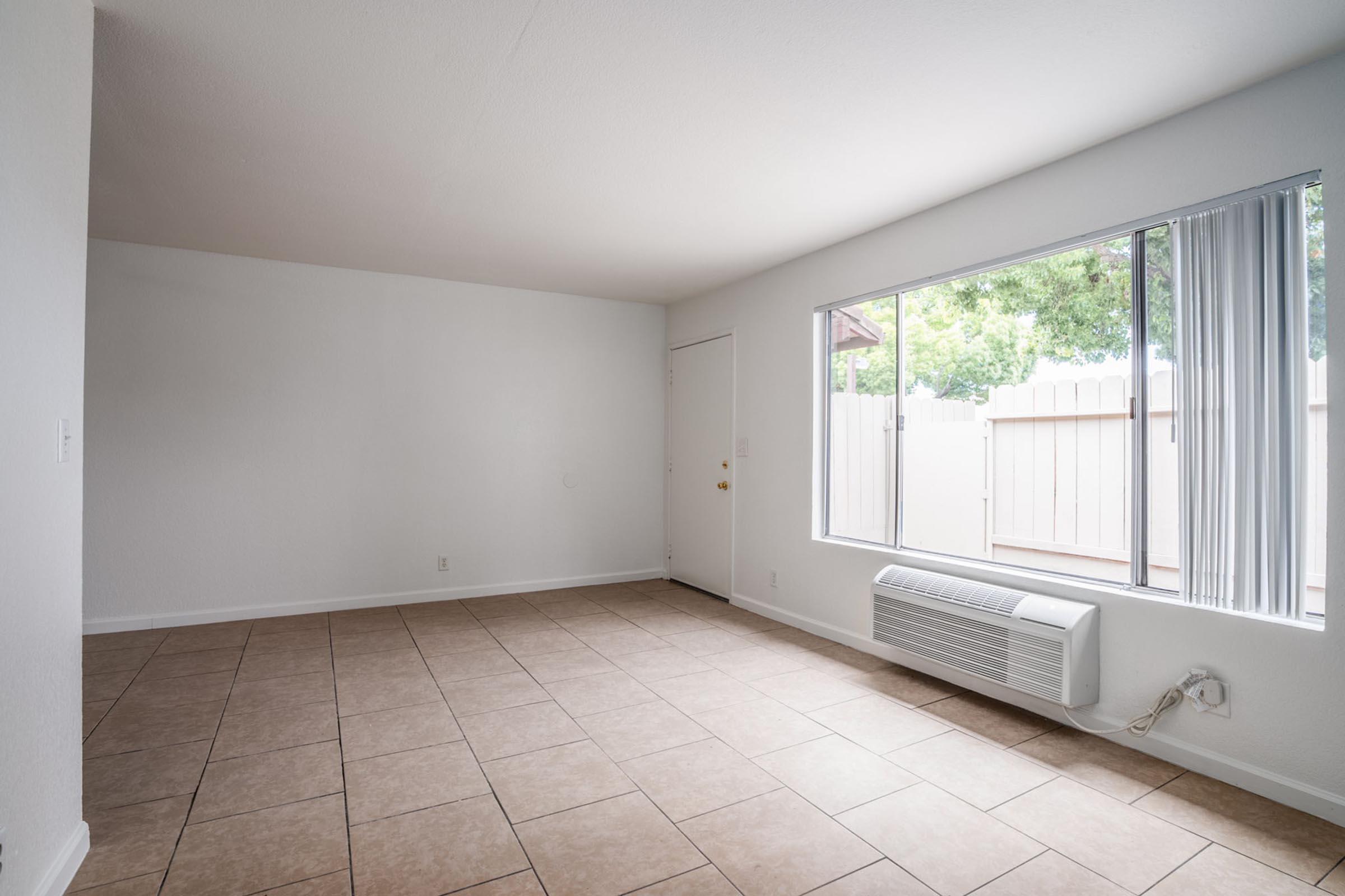 A spacious, empty room with beige tile flooring, white walls, and a large window allowing natural light. There is an air conditioning unit beneath the window, and a door to the left leading outside. The room features a simple layout with minimal furnishings, creating a bright and open atmosphere.