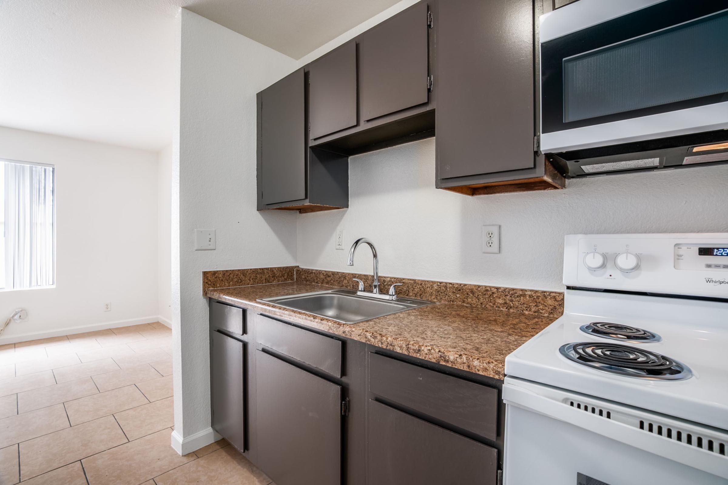 A kitchen area featuring dark wood cabinets, a countertop with a sink, and a white stove and microwave. The floor is tiled and there is a window illuminating the space. The walls are painted in a light color, creating a clean and minimalist look.