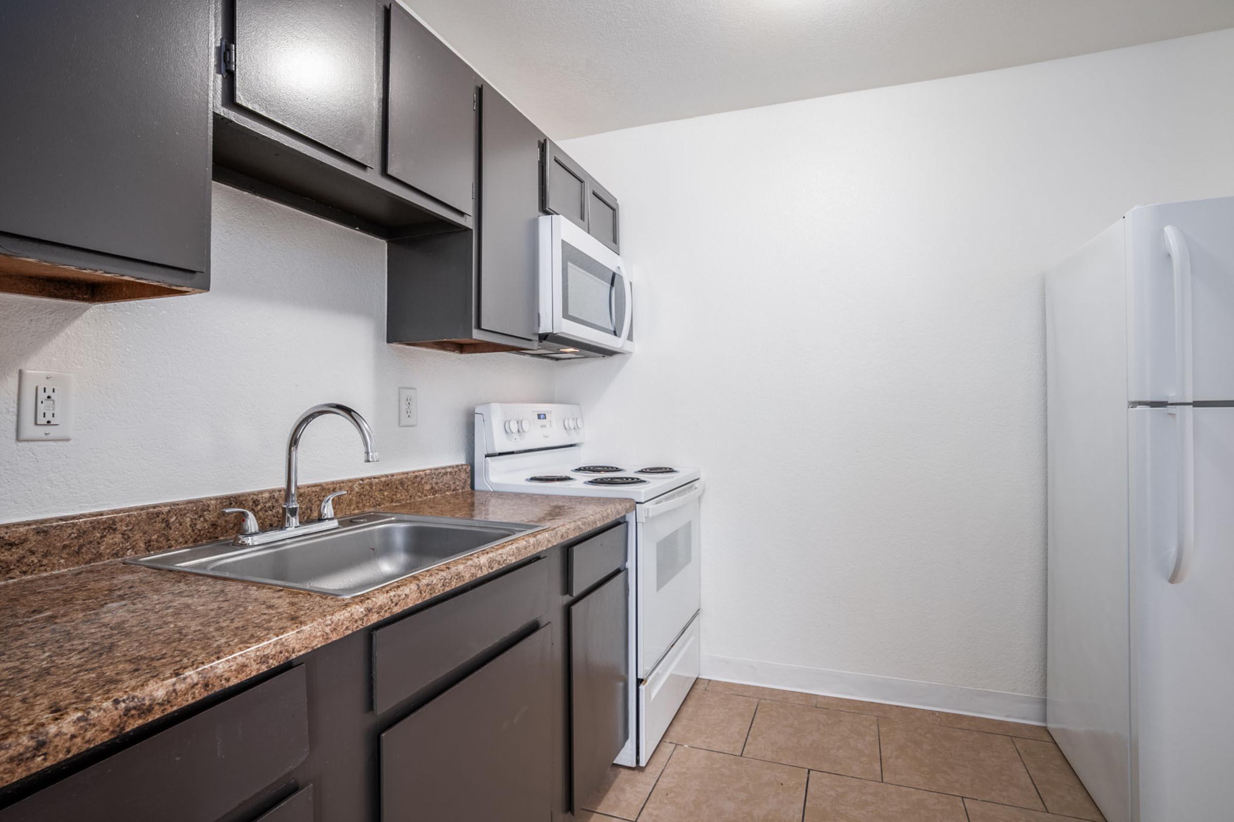 A small kitchen featuring dark cabinets, a stainless steel sink, a white stove, and a microwave above it. There's a white refrigerator in the corner and a granite countertop. The walls are painted white, and the floor consists of square tiles. The overall atmosphere is clean and modern.