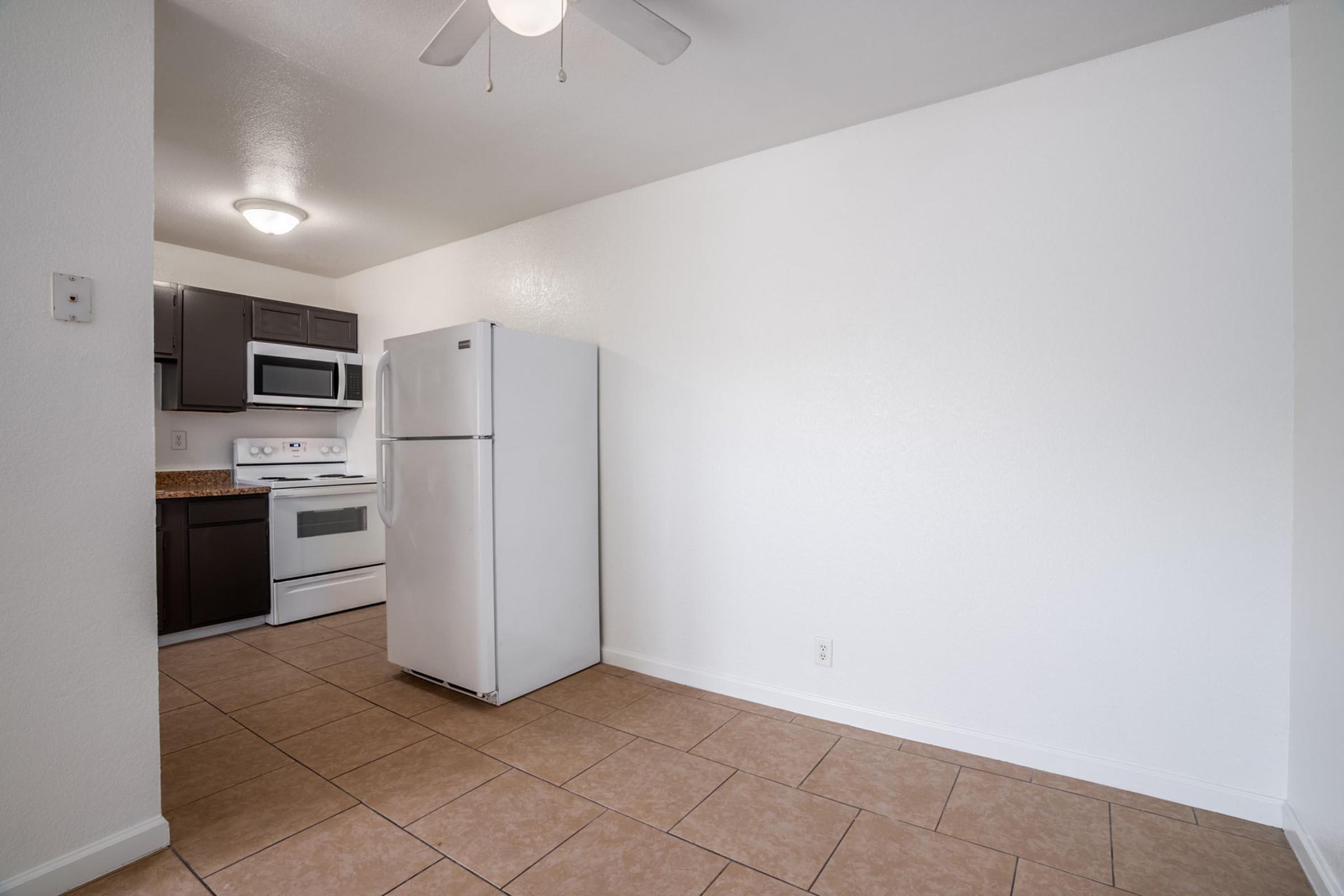 A clean, empty kitchen space featuring light-colored walls, tile flooring, and a ceiling fan. The kitchen includes a white refrigerator, an oven, and dark cabinetry. Natural light illuminates the area, creating a bright and inviting atmosphere.