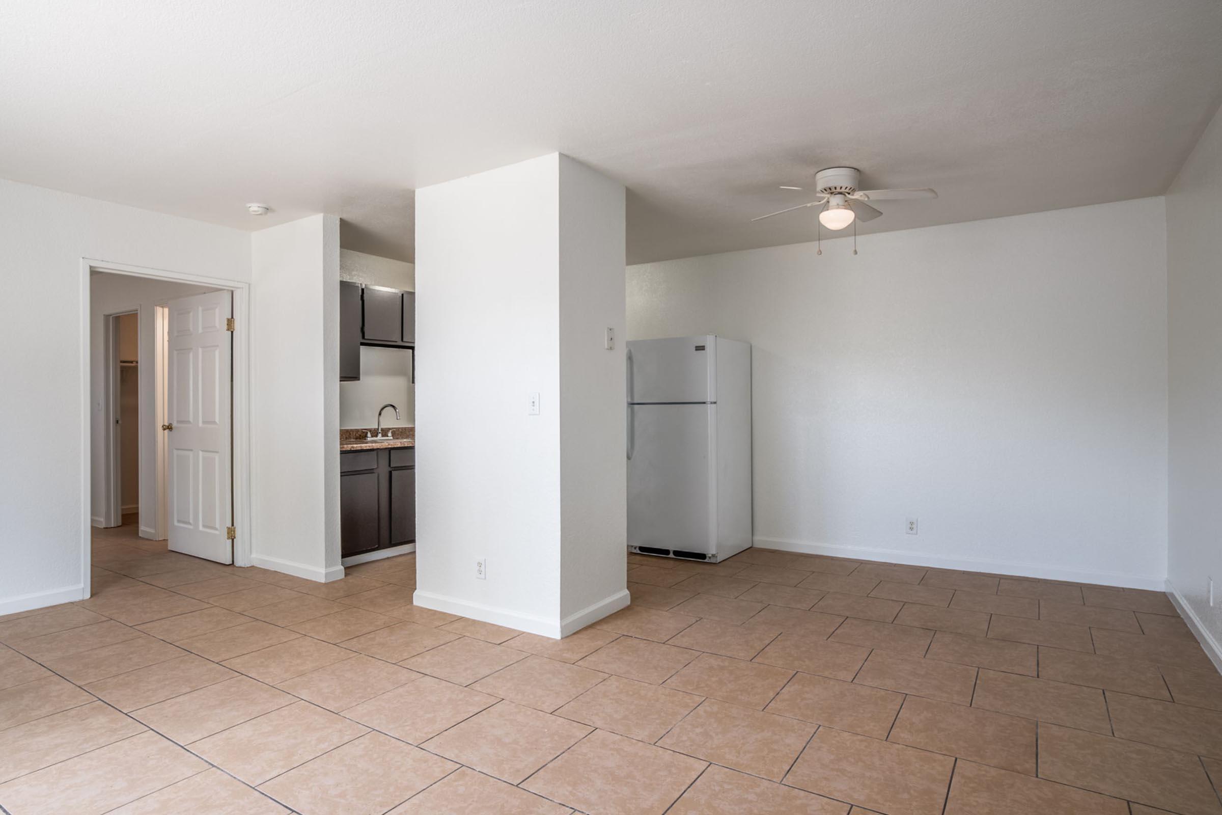 A bright, empty living space featuring beige tiled flooring, a ceiling fan, and a small kitchen area with dark cabinets. A white refrigerator stands against the wall. A door leads to another room, indicating an open layout. The walls are painted in a light color, enhancing the airy feel of the room.