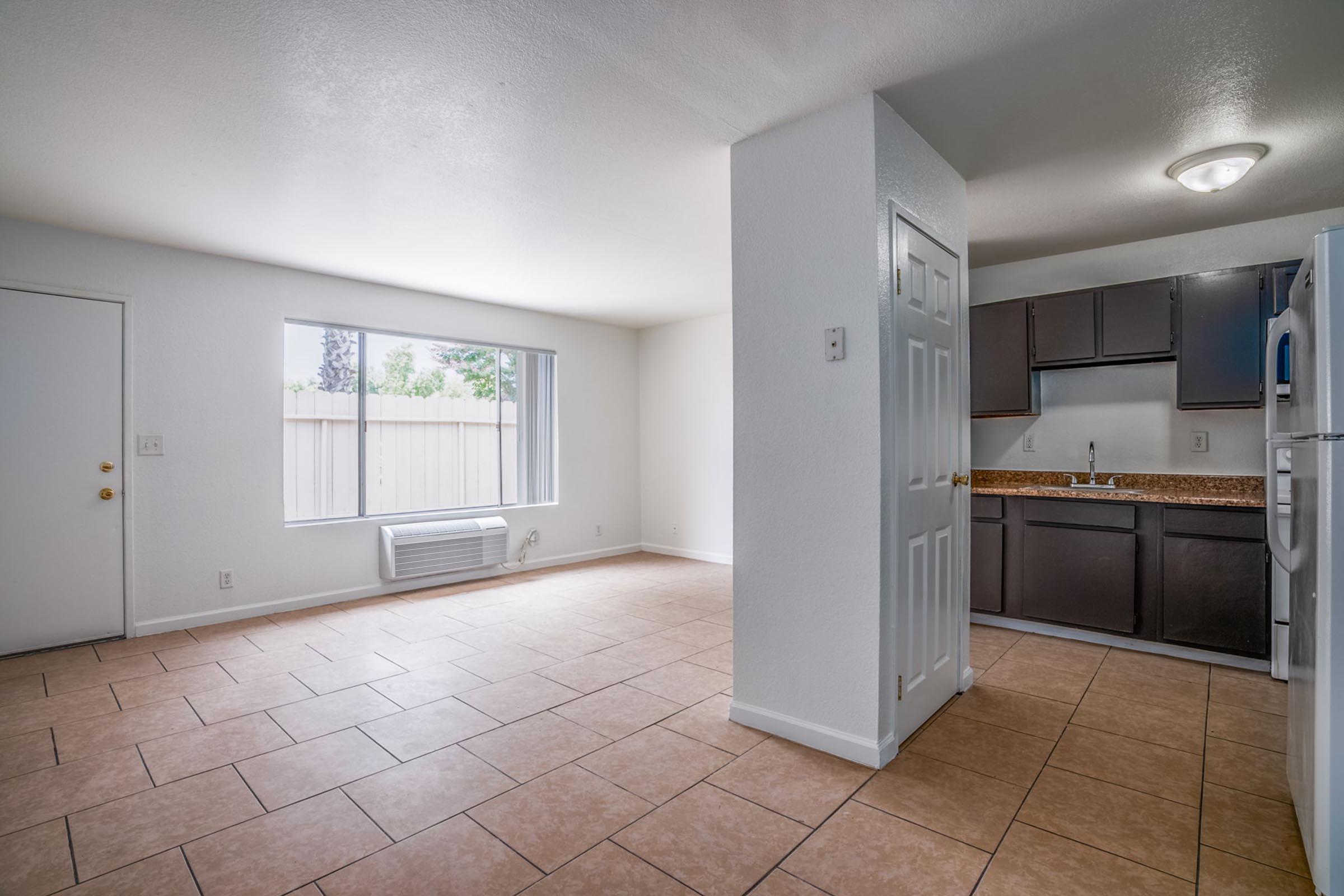 Interior view of a spacious, well-lit room featuring a tiled floor. To the left, there is a large window allowing natural light, and on the right, a kitchen area with dark wooden cabinets and a countertop. The room is painted in light colors, creating an open and inviting atmosphere.