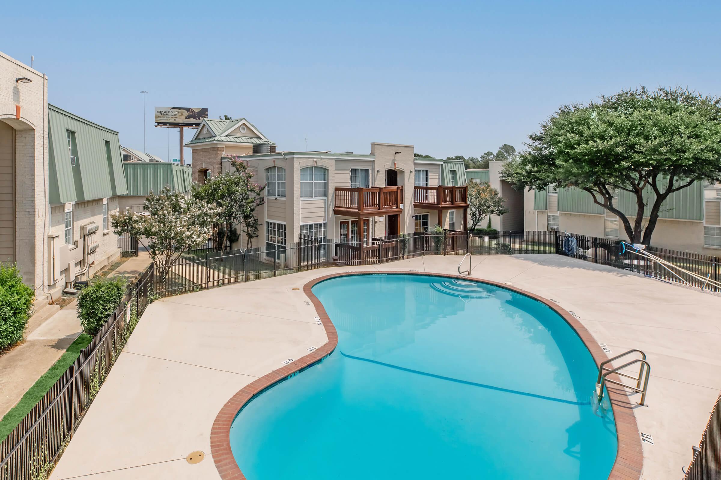 View of a residential pool area with a clear, blue swimming pool surrounded by a fence. In the background, there are two buildings with balconies, trees, and a billboard visible. The sky is bright and clear, suggesting a sunny day.