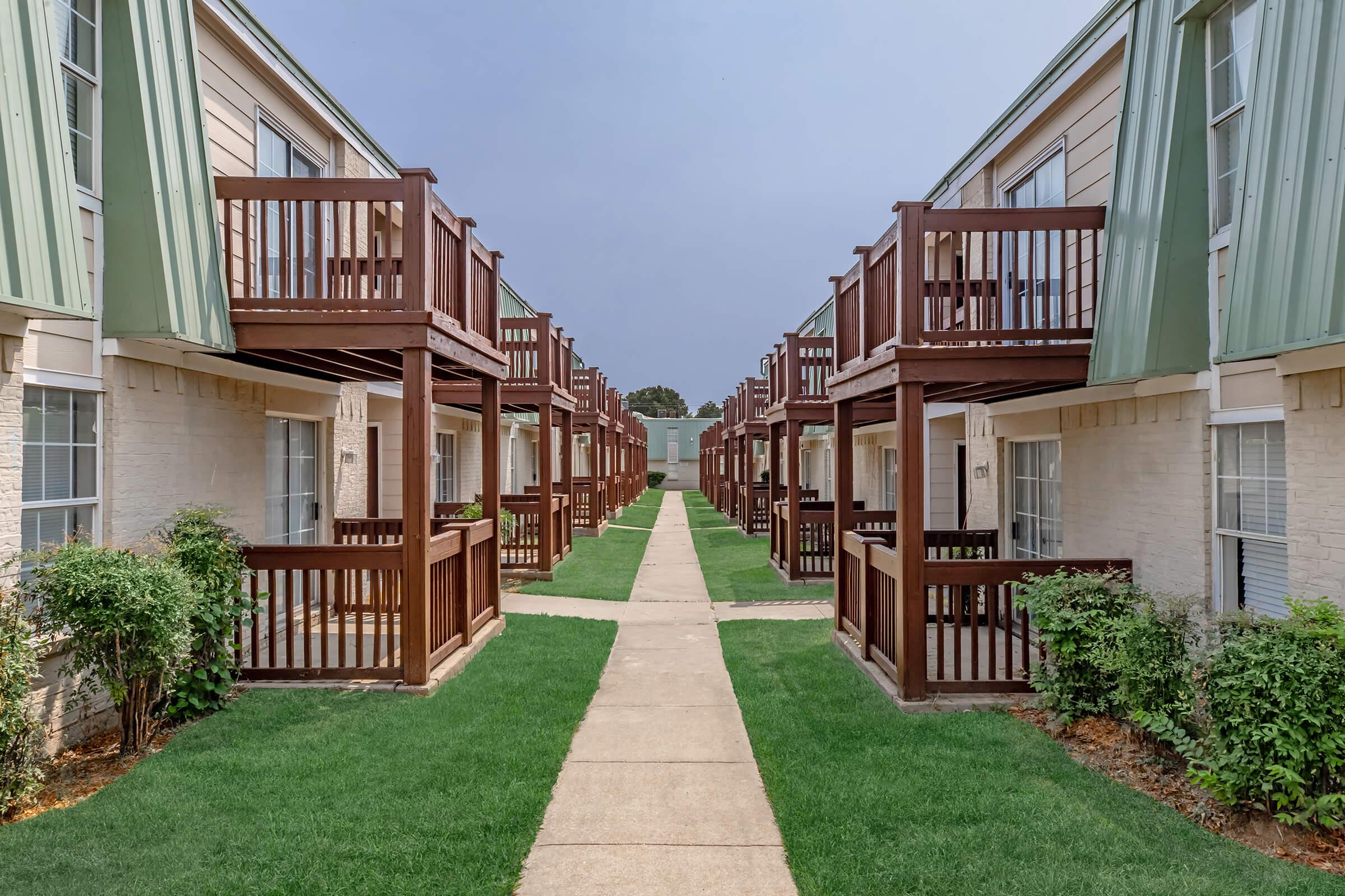 A view of a courtyard with two-story apartment buildings on either side. The buildings have balconies made of wood, and there is a well-maintained path and green grass in the center. The sky is slightly overcast, creating a calm and serene atmosphere.
