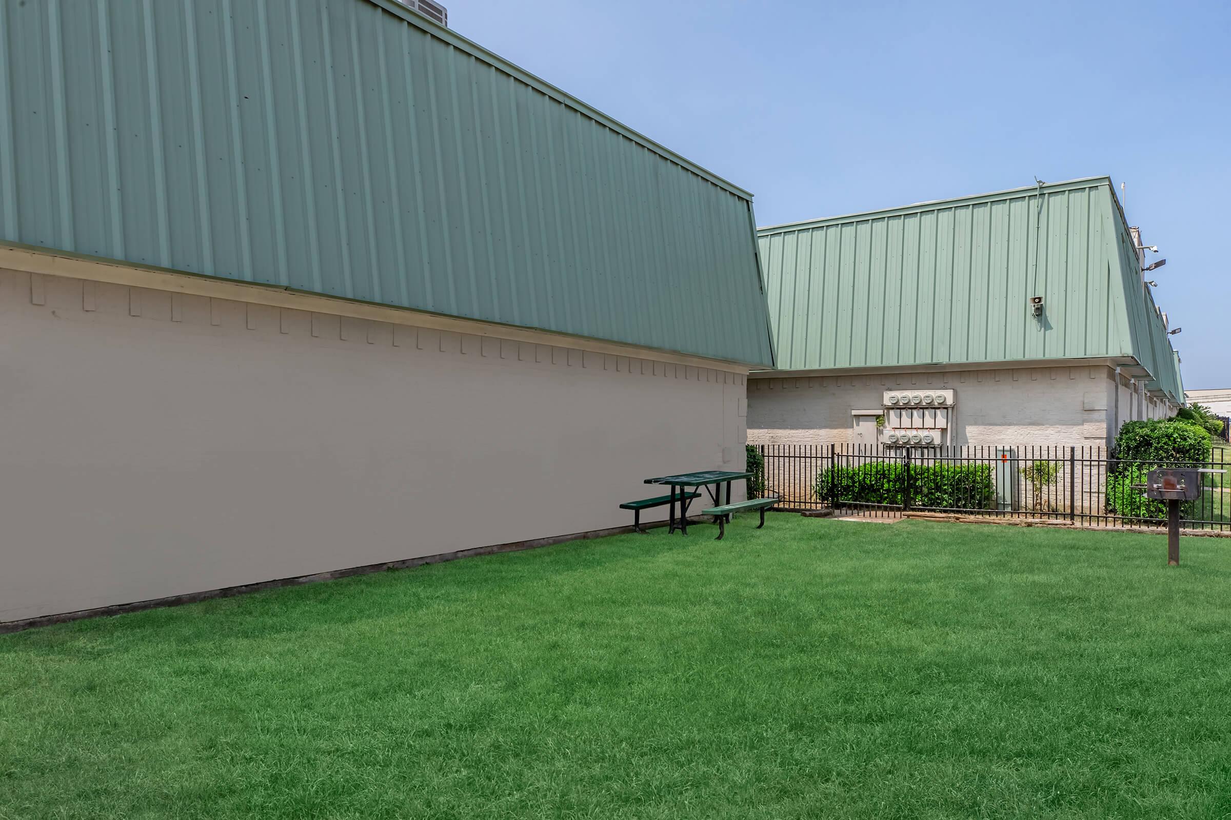 A grassy area featuring a picnic table, surrounded by two buildings with green roofs. The setting is clear and sunny, with a fence and some shrubs in the background.
