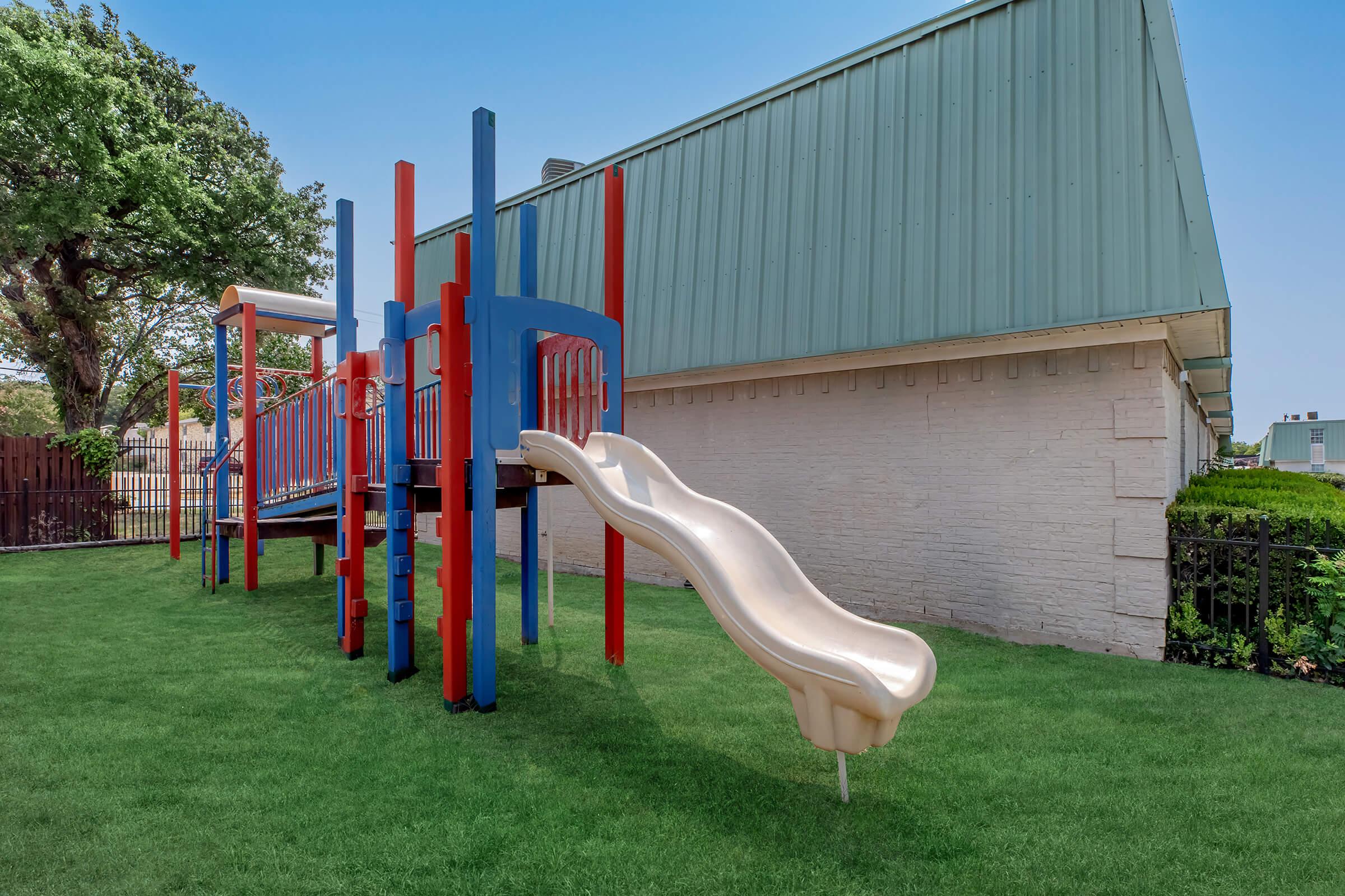 A colorful playground featuring a slide and climbing structure, set on well-maintained grass. The playground has red and blue elements, with a backdrop of a green roofed building and trees providing shade in the background. Ideal for children to play and socialize.