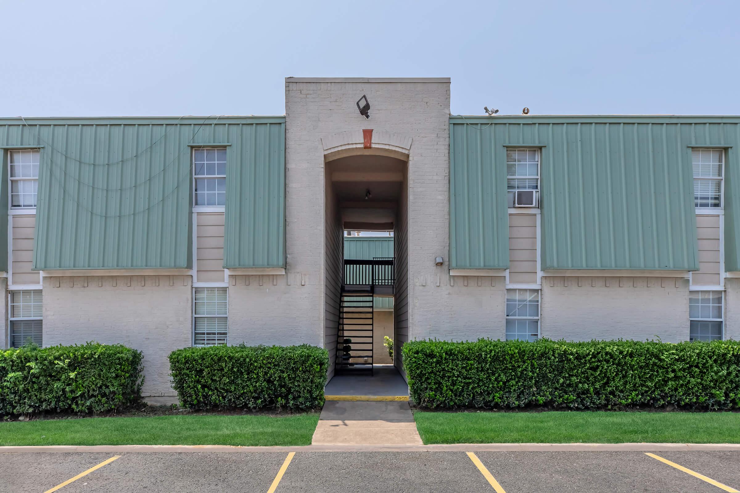 A two-story apartment building with a light-colored brick exterior and a green metal roof. The entrance features a covered walkway and stairs leading to the second floor. Neatly trimmed hedges line the front, and parking spaces are visible in the foreground on a paved area.